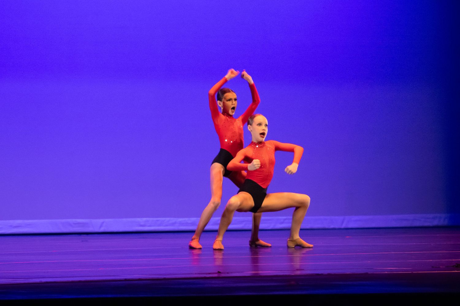 Two dancers in red costumes perform a dramatic contemporary dance routine on a blue-lit stage.
