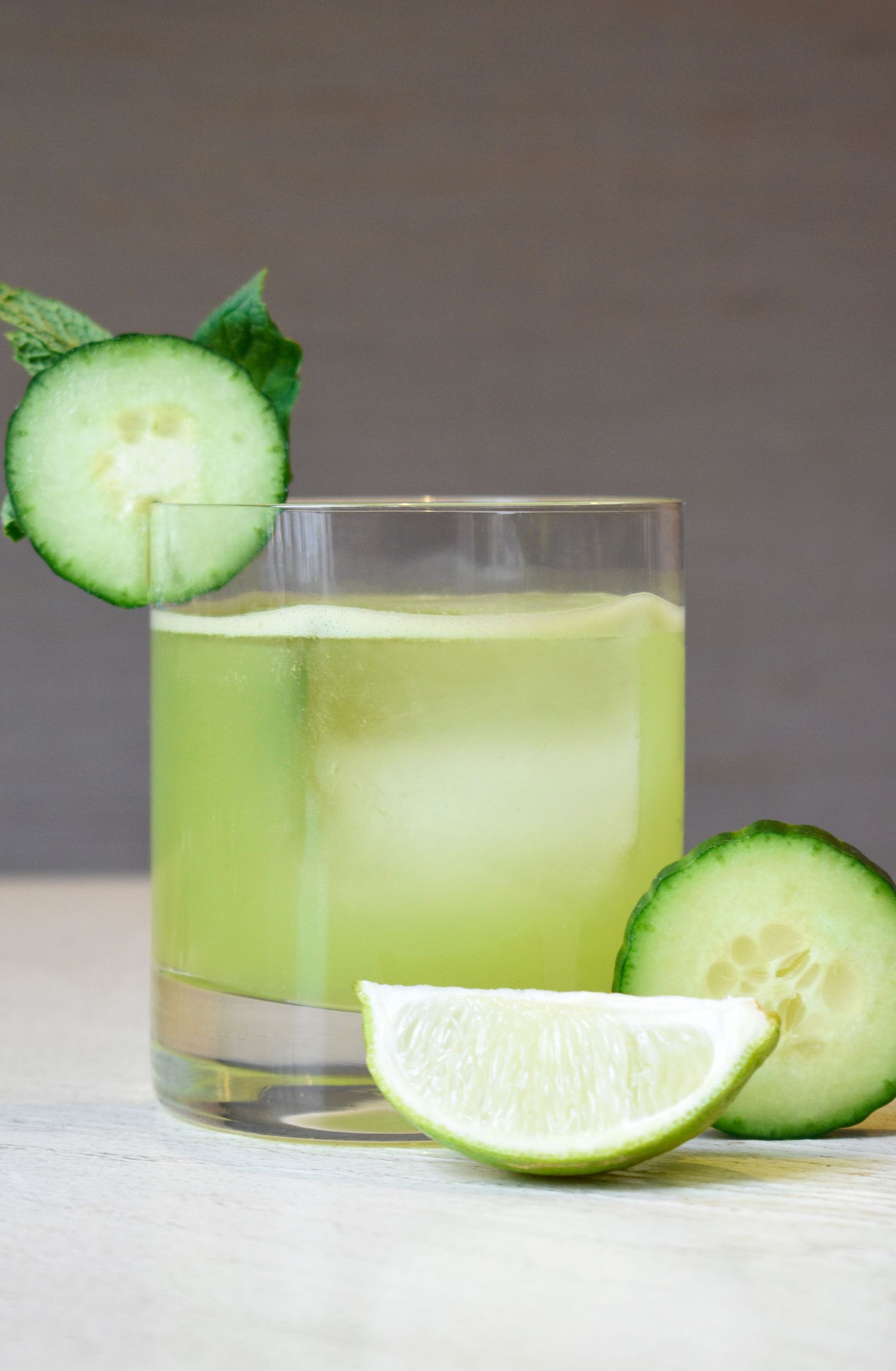 A bright green cucumber lime cocktail with ice, cucumber slice, and mint garnish on a white surface.
