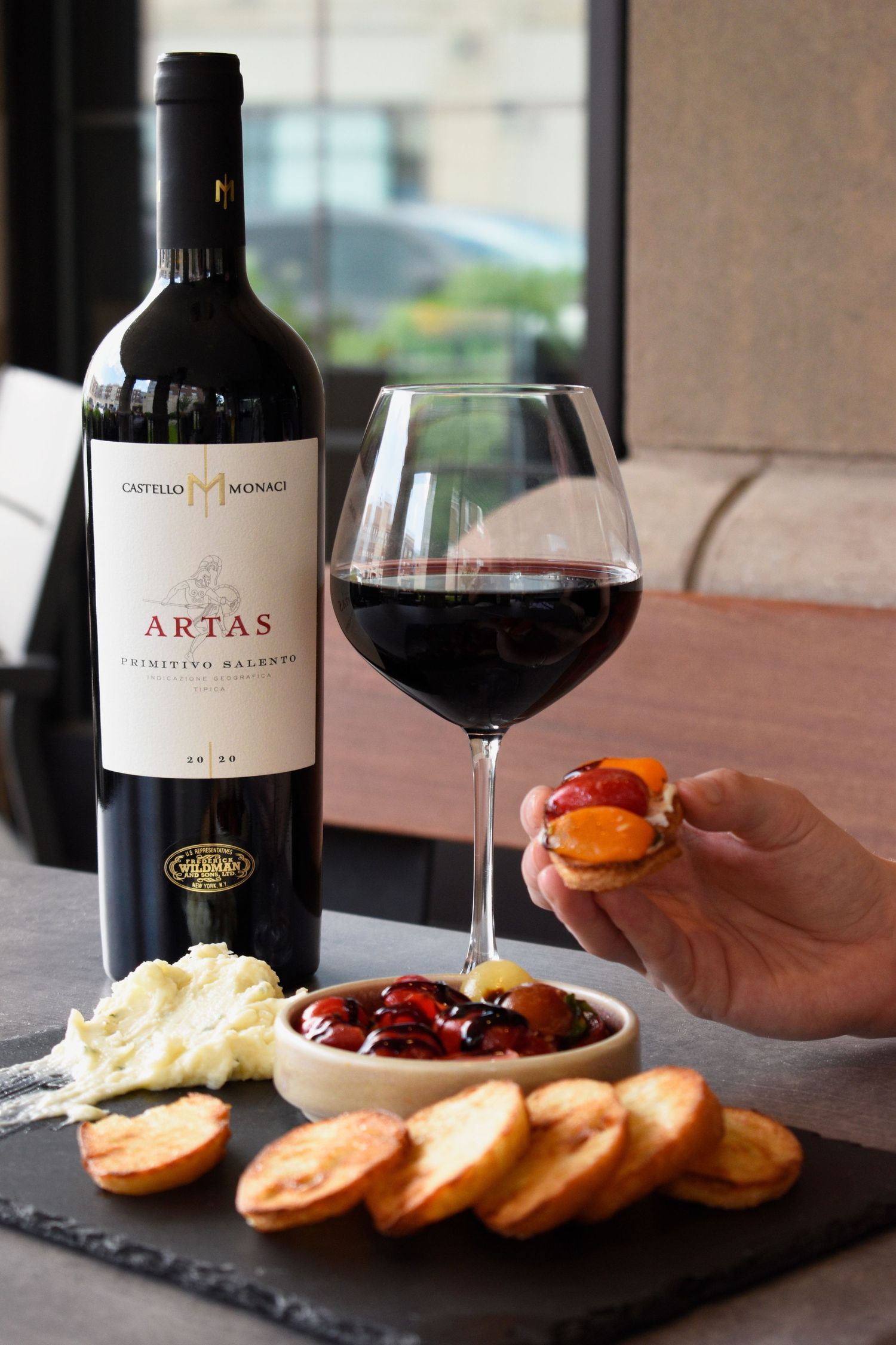 Castello Monaci Artas Primitivo Salento red wine bottle with glass and bruschetta appetizers on slate board.