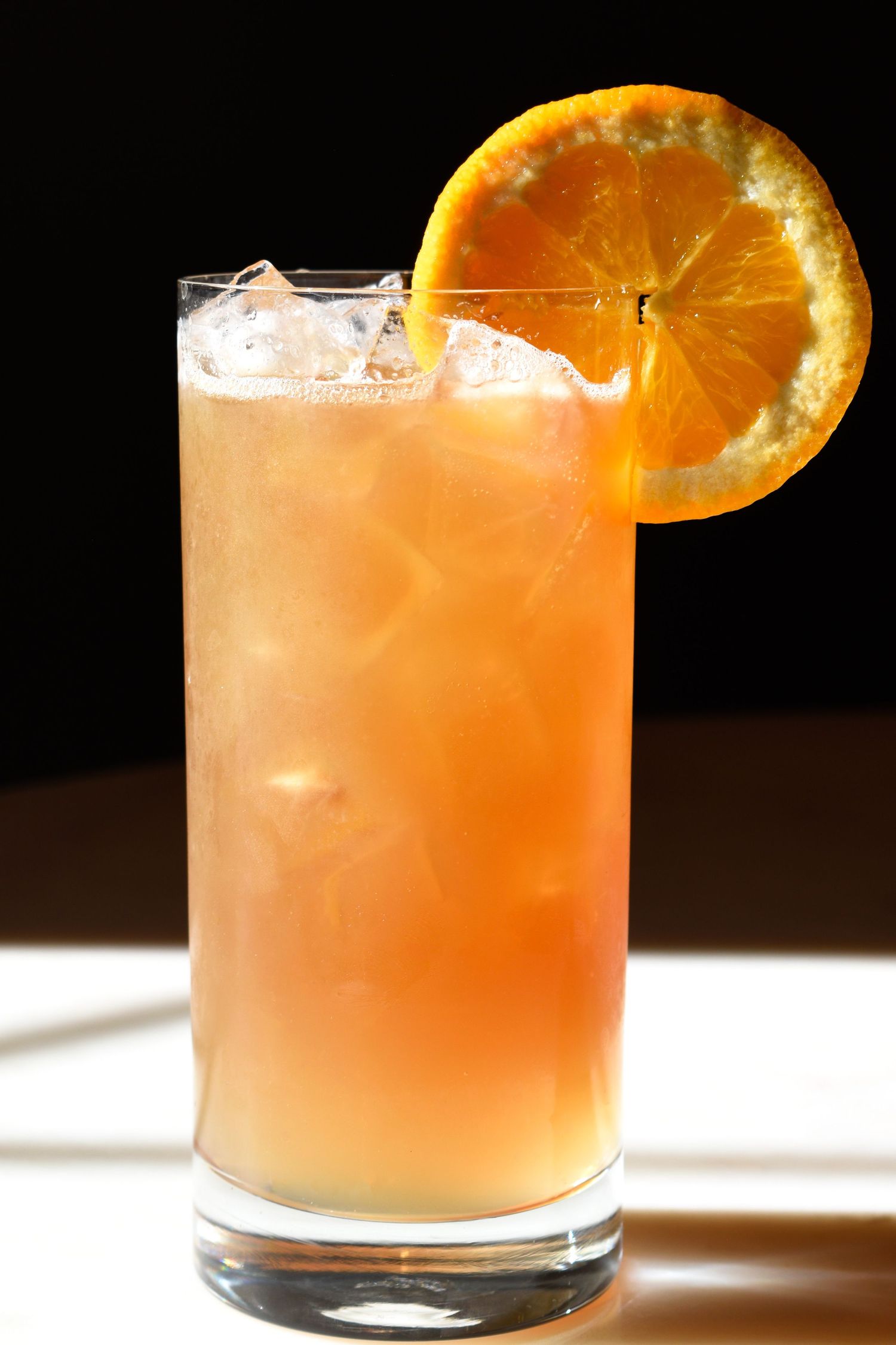 A tall highball glass with an orange-hued iced cocktail garnished with a fresh orange wheel slice.