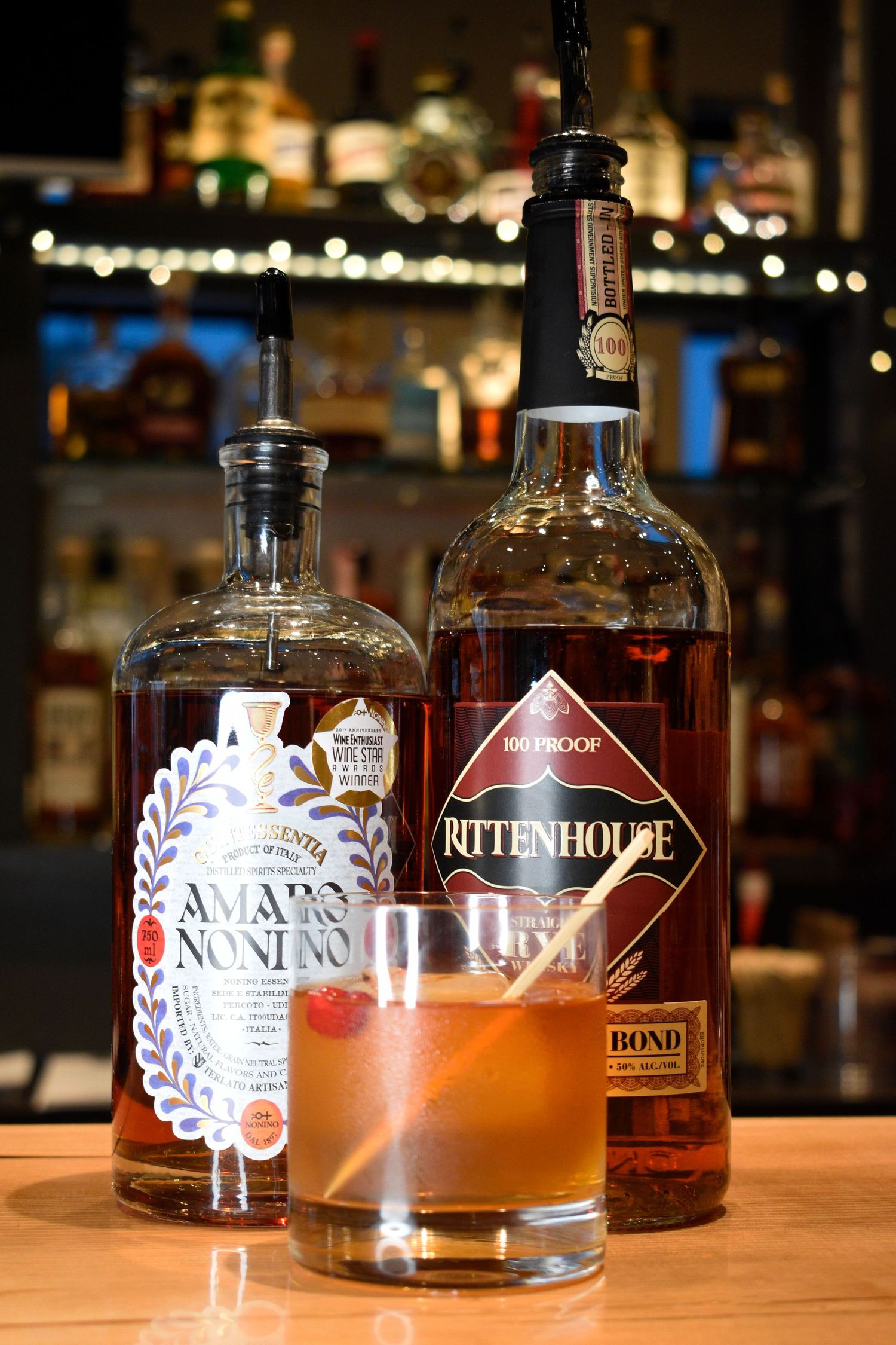 A cocktail glass with Amaro Nonino and Rittenhouse 100 Proof Rye whiskey bottles displayed on a bar.