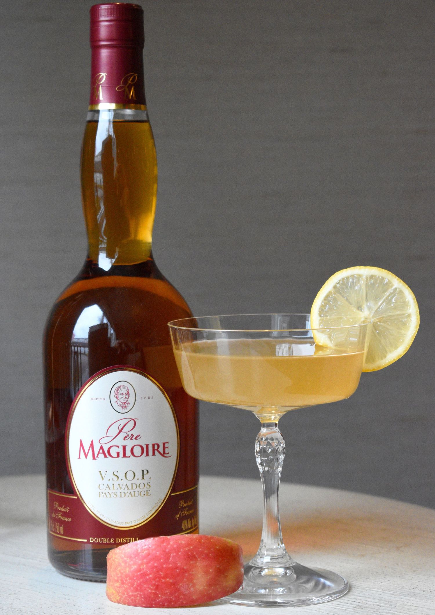 Golden Calvados cocktail in a coupe glass with lemon garnish beside a Pere Magloire VSOP bottle and peach slice.