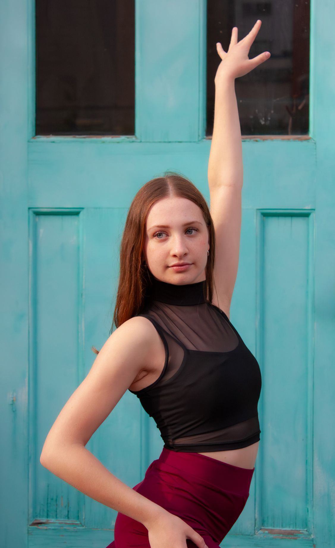 Dancer in black mesh crop top raises arm gracefully in front of turquoise double doors.