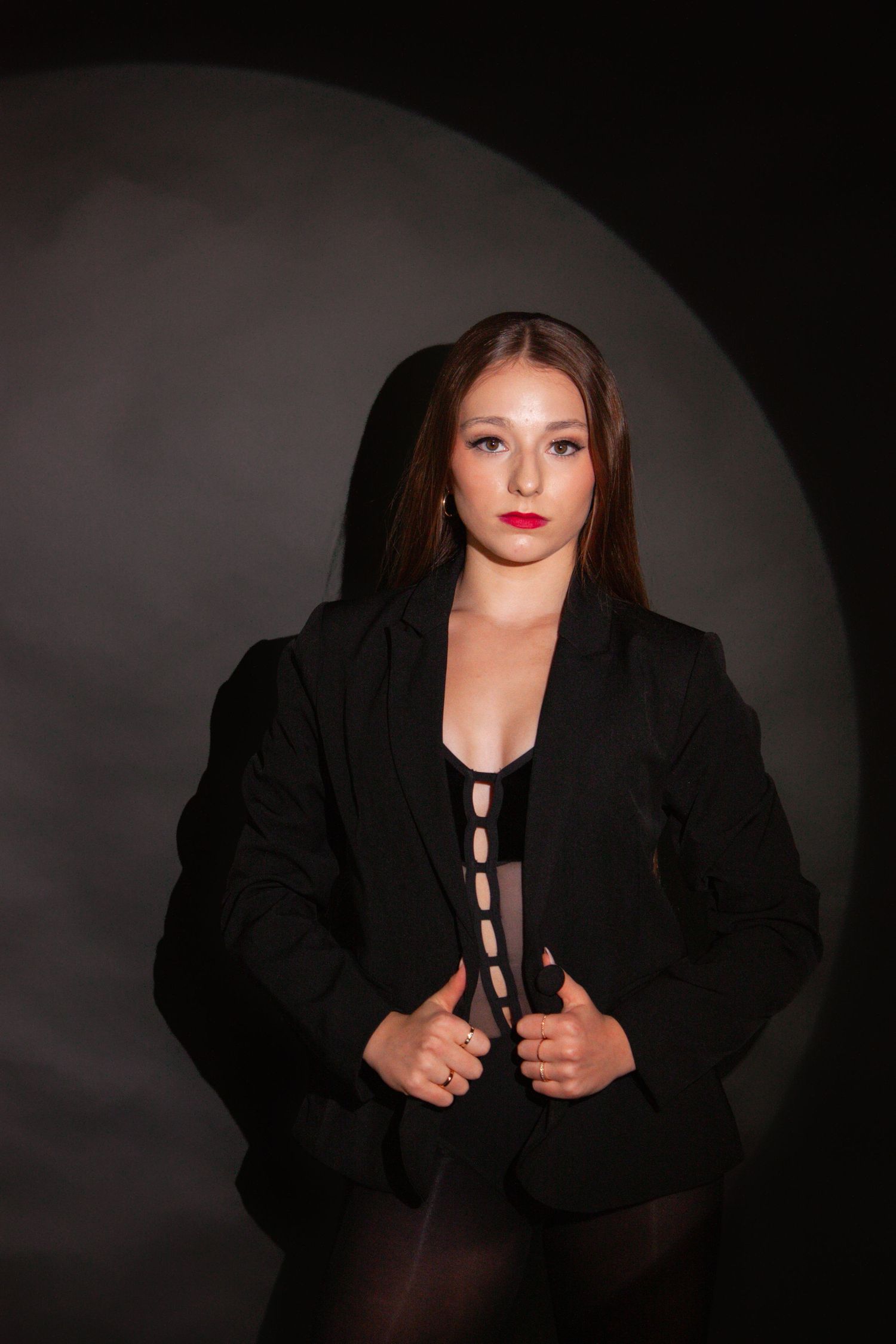 Young woman in a black blazer and bodysuit posing confidently in dramatic studio lighting against a dark backdrop.