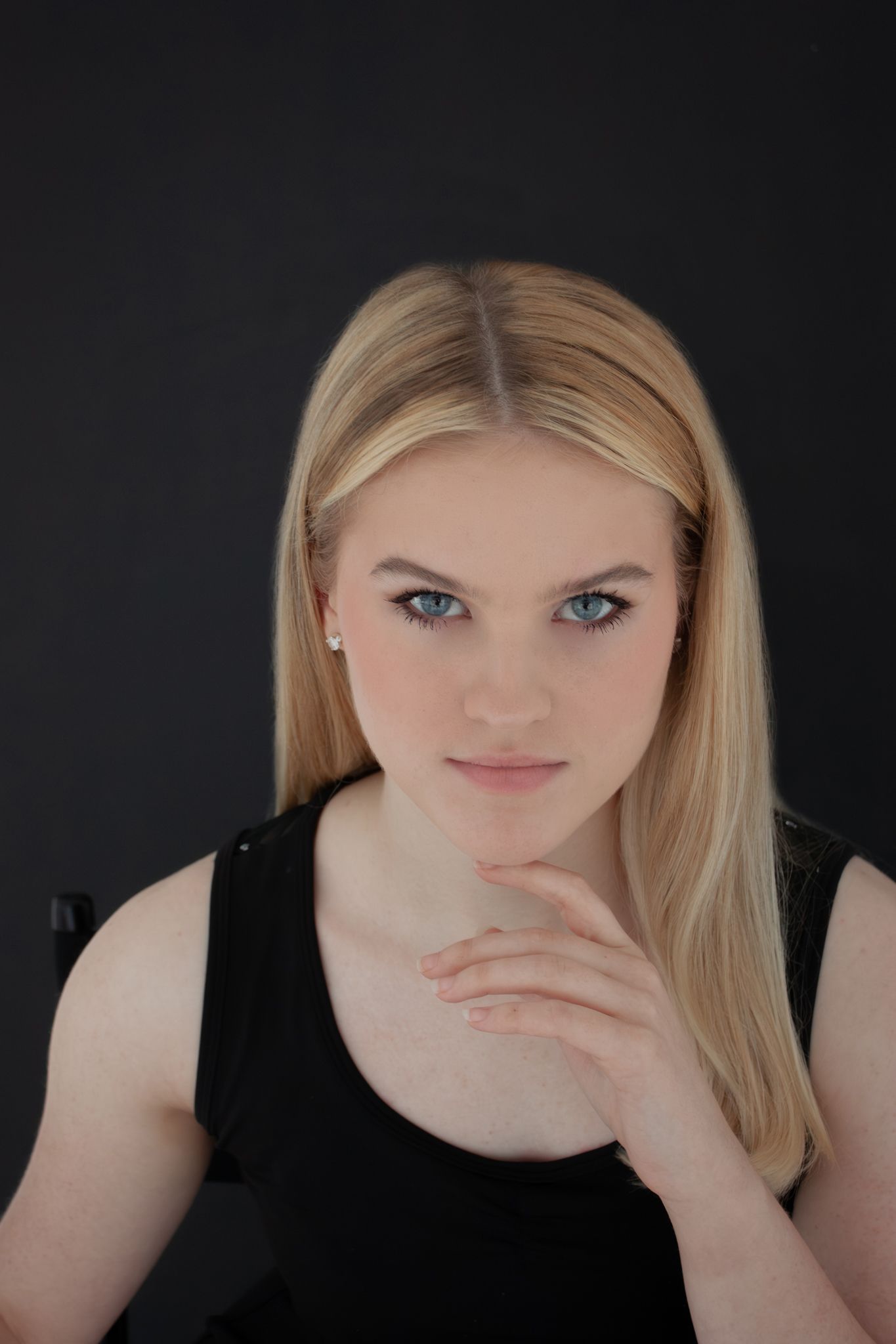 Blonde woman with blue eyes in black top rests chin on hand against a dark background.