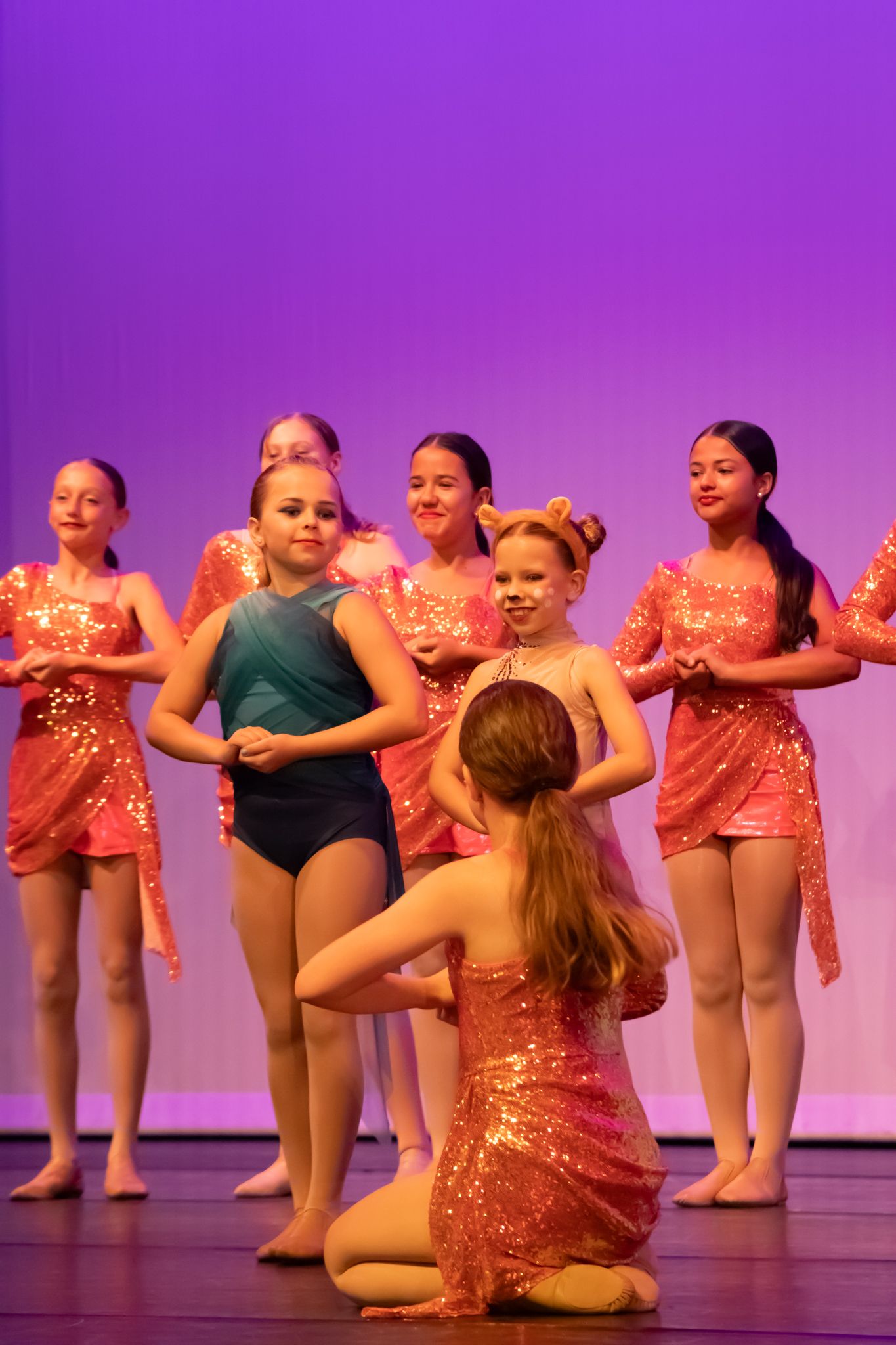 Girls in sparkly orange costumes perform a jazz dance routine under purple stage lights at a recital.