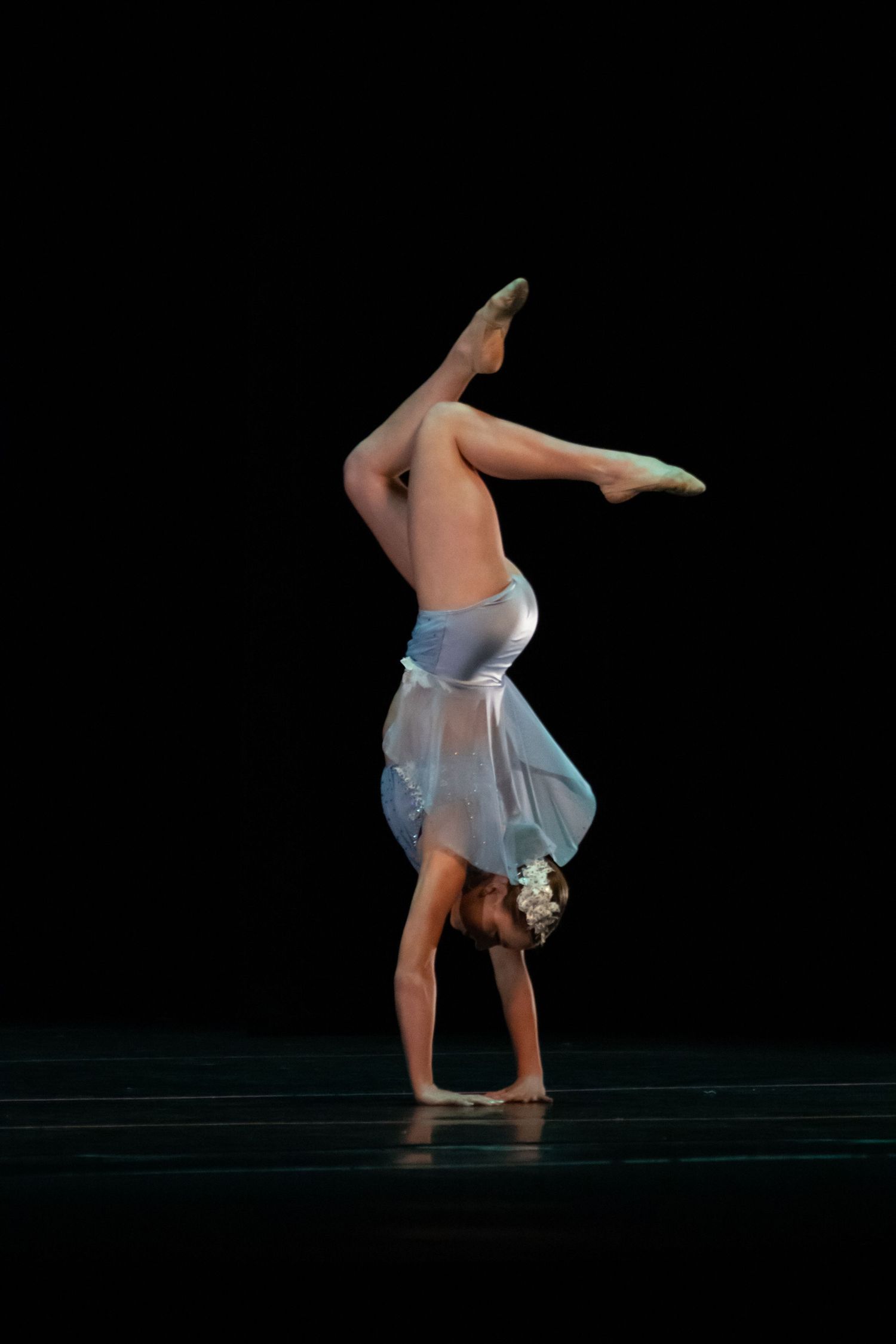 A skilled dancer performs a headstand with legs split on a dark stage wearing a silver and blue costume.
