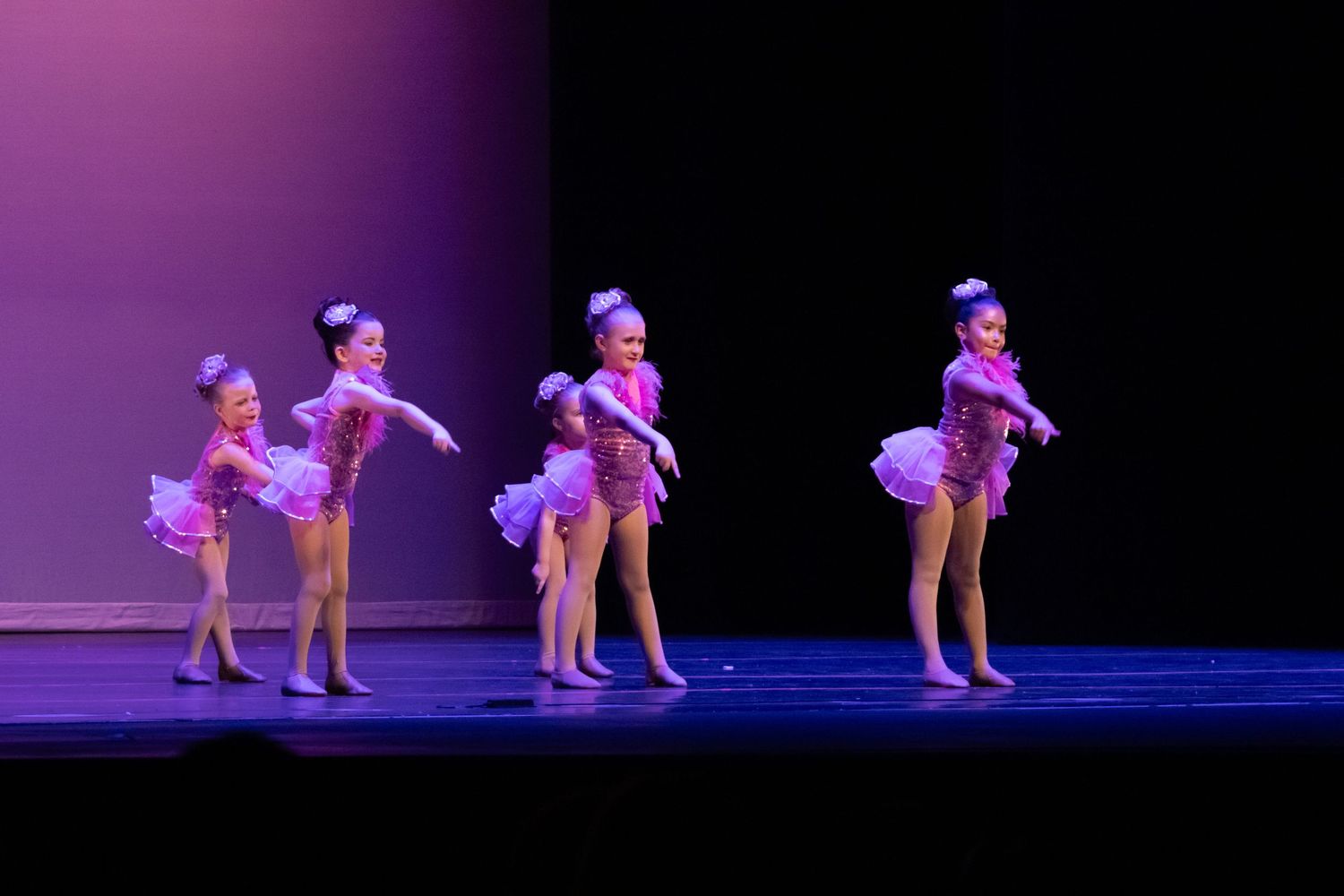 Four young girls in pink sequin costumes perform a synchronized dance routine on stage.