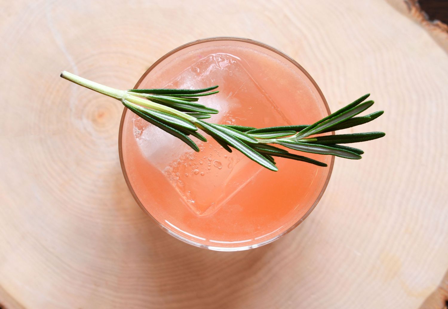 A refreshing pink cocktail with ice and a fresh rosemary sprig garnish served in a rocks glass on a wood slice.