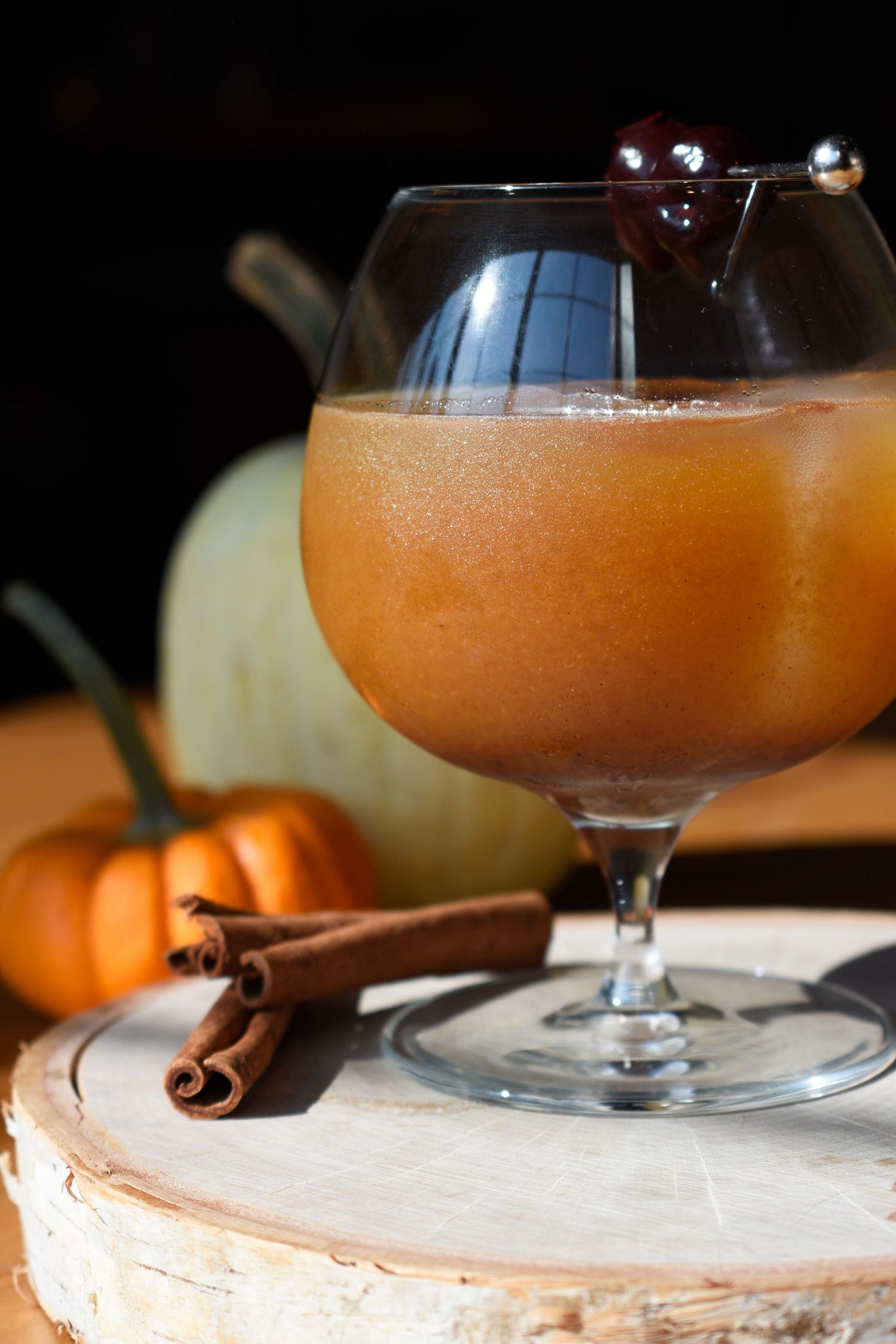 Pumpkin spice cocktail in a brandy snifter with cinnamon sticks and decorative pumpkins on a rustic wood base.