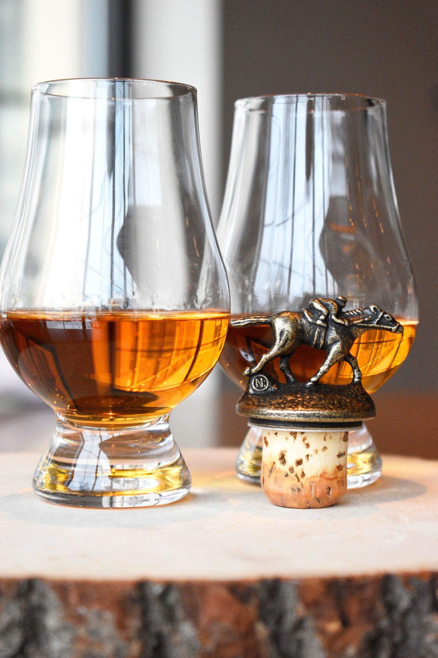 Two Glencairn glasses of bourbon whiskey with Blanton's horse and jockey stopper on a rustic wood stump.