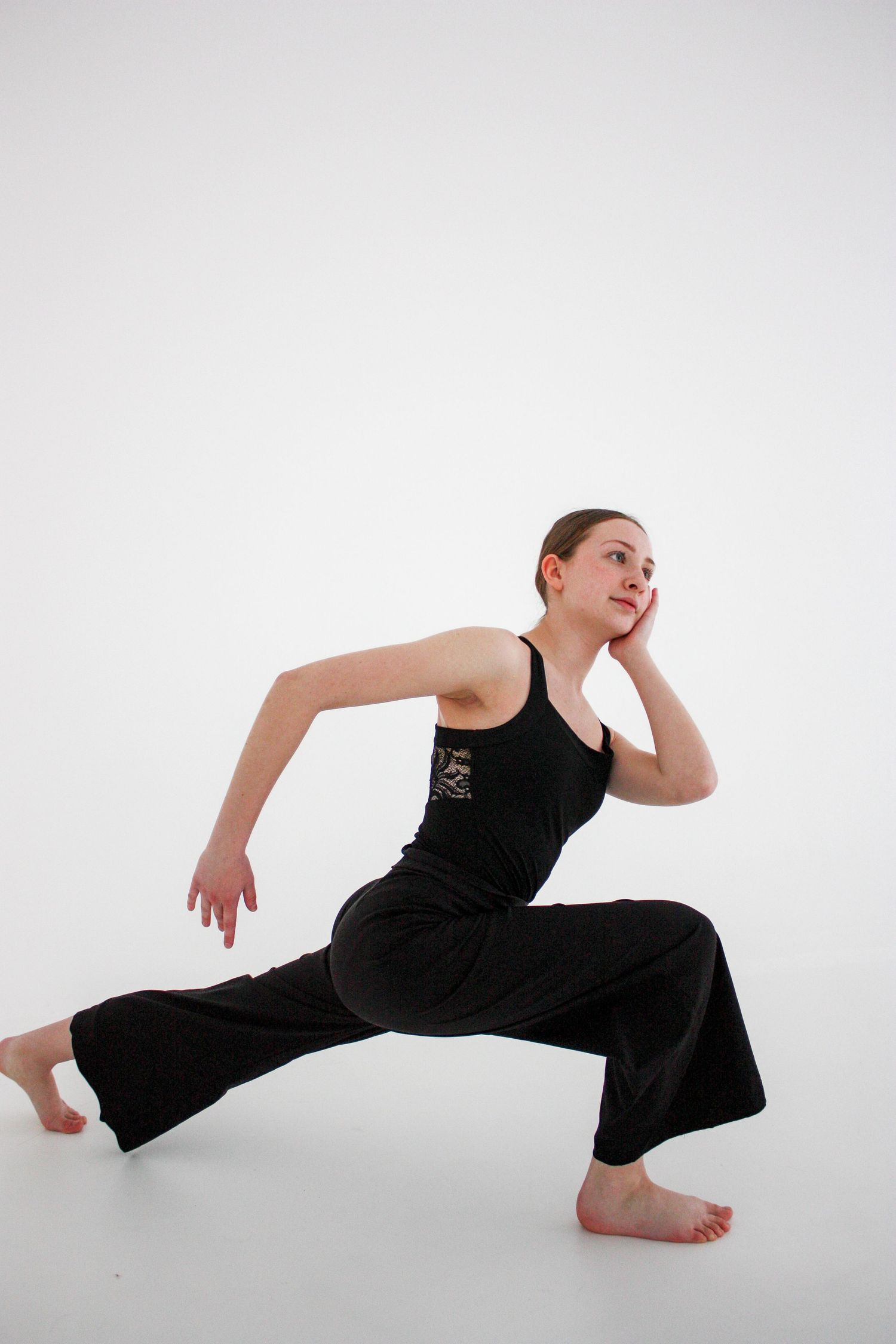 A young dancer in black wide-leg pants performs a deep lunge pose barefoot against a white background.