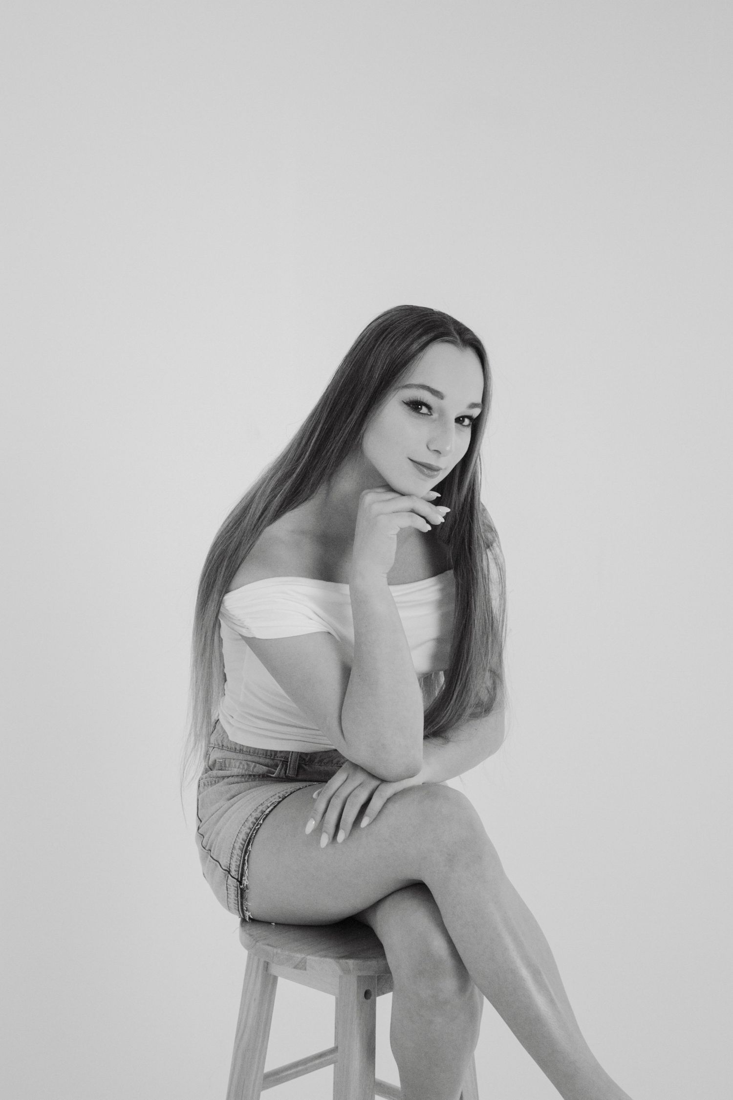 Young woman in white off-shoulder top and denim skirt sits on stool in black and white studio portrait.