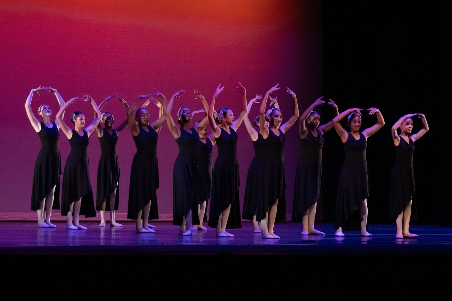 A line of dancers in black dresses perform with arms raised on a colorful stage.