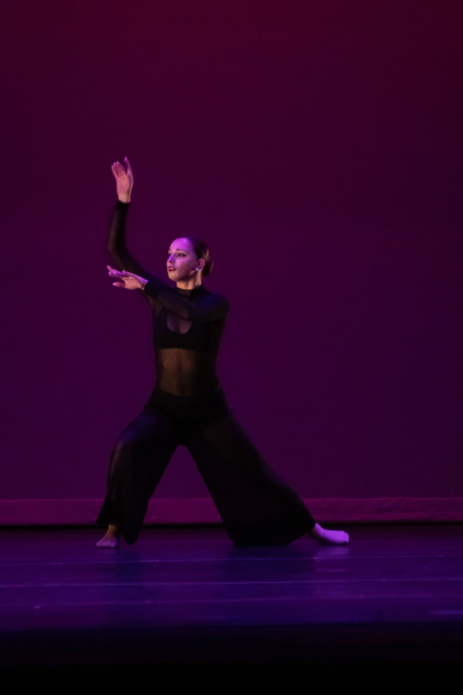 A contemporary dancer in all-black performs an expressive pose on a purple-lit stage.