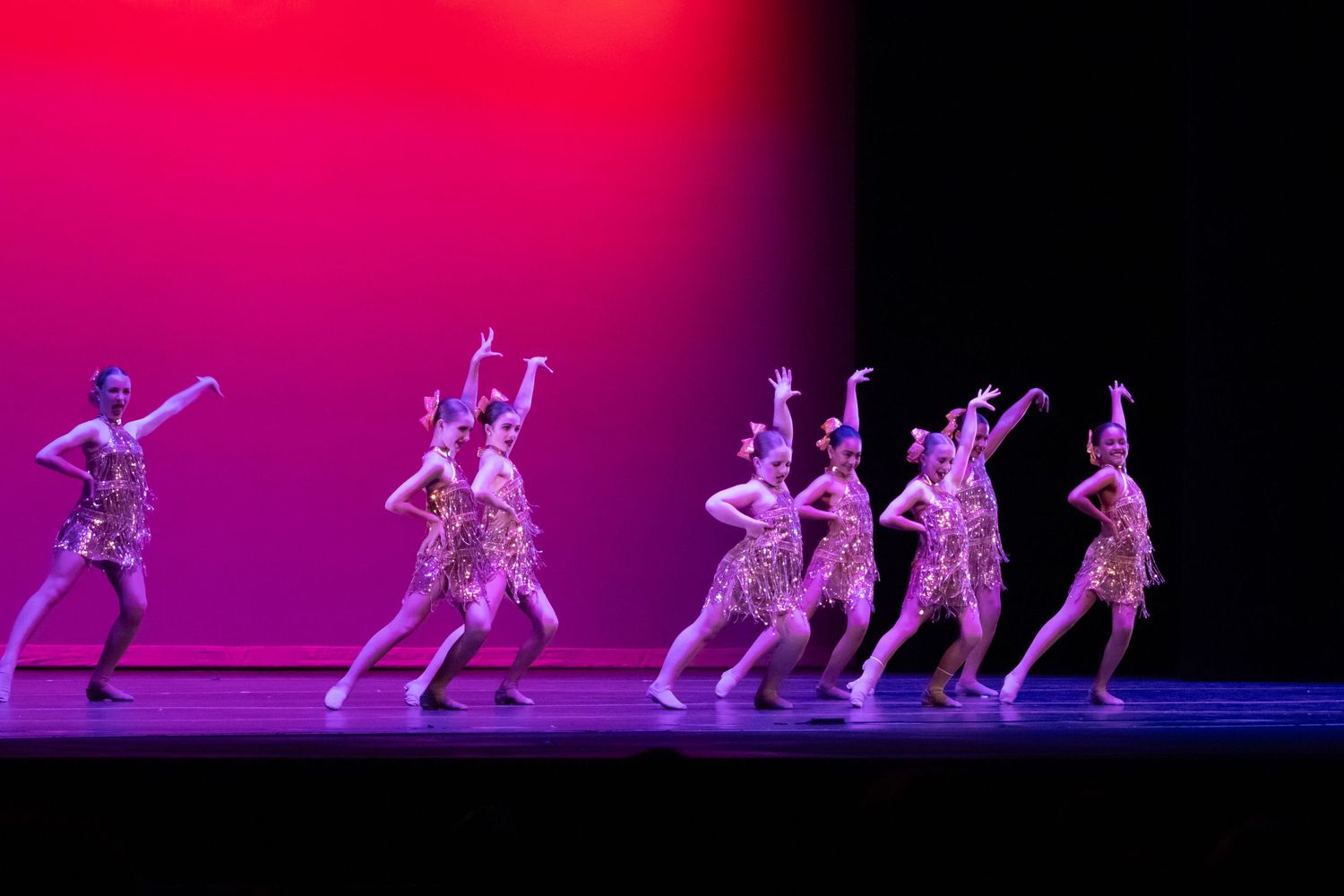 A line of dancers in gold sequined costumes perform in sync on a pink and purple-lit stage.