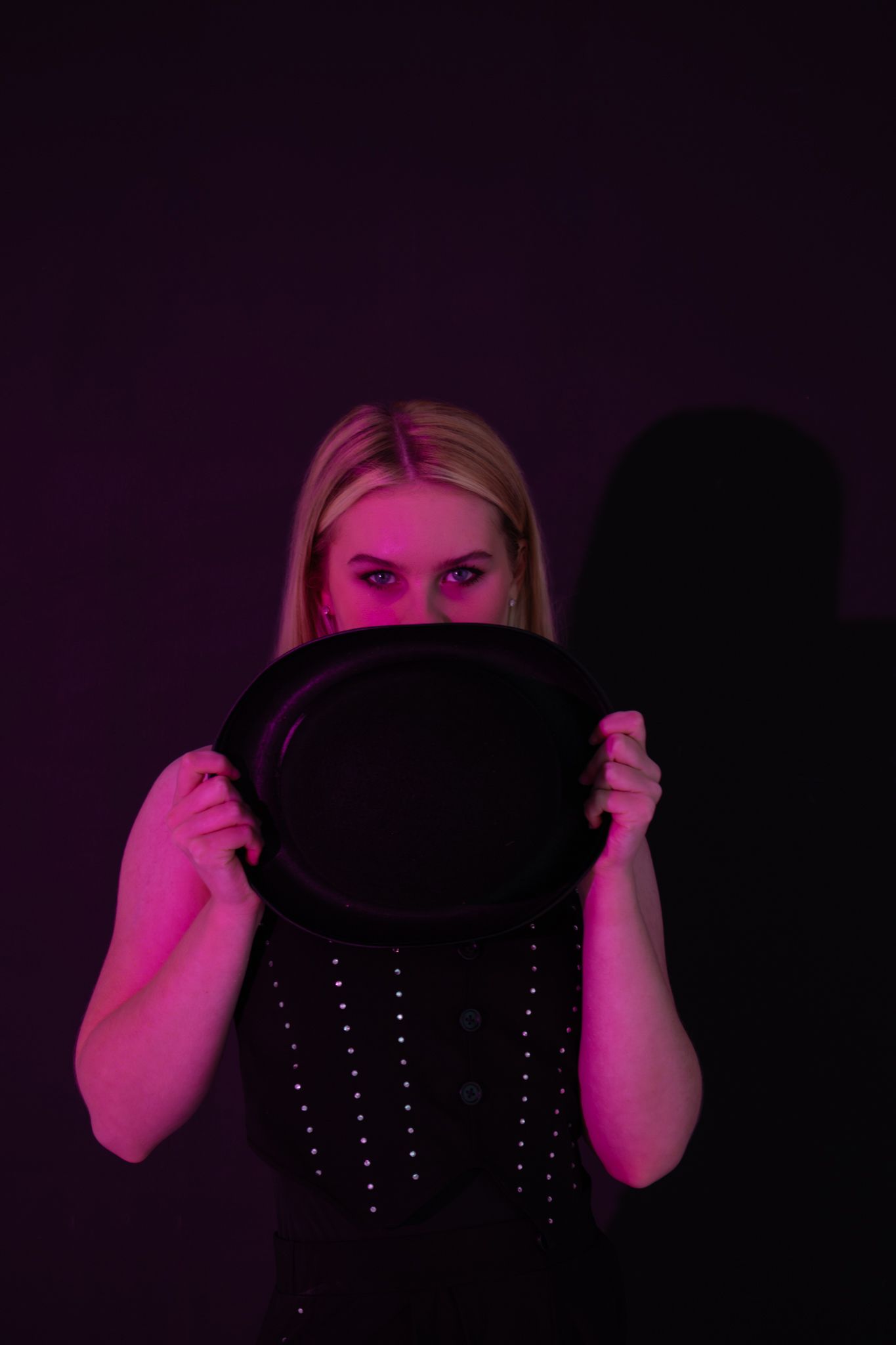 Blonde woman hiding face behind black bowler hat under pink neon light.