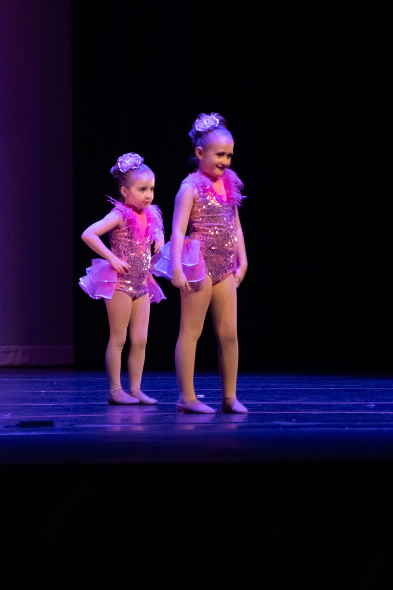 Two young girls perform on stage in sparkly pink sequin dance costumes with tutus and tiaras under stage lighting.