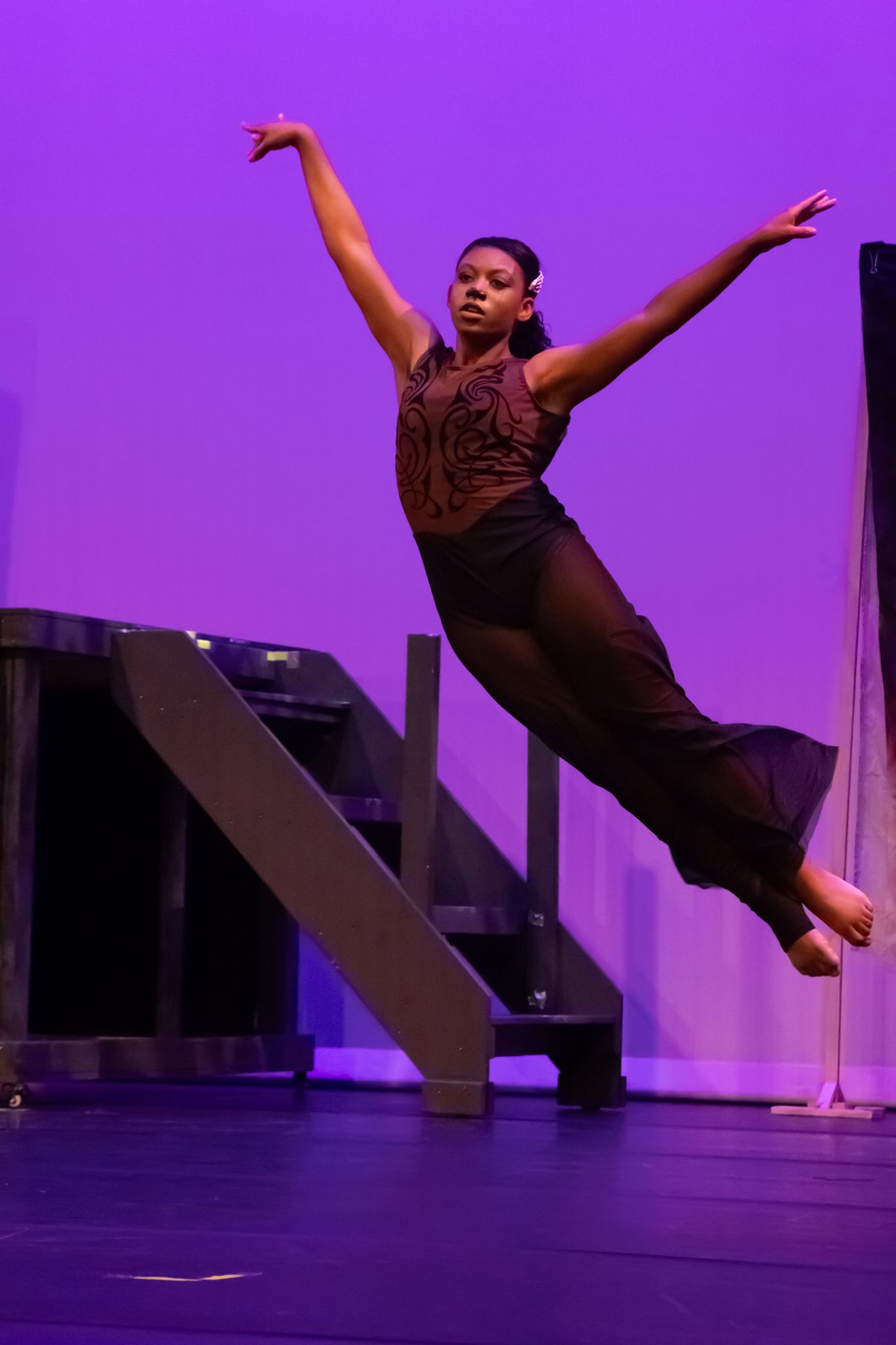 Young Black female dancer leaps gracefully in the air under purple stage lighting during recital.