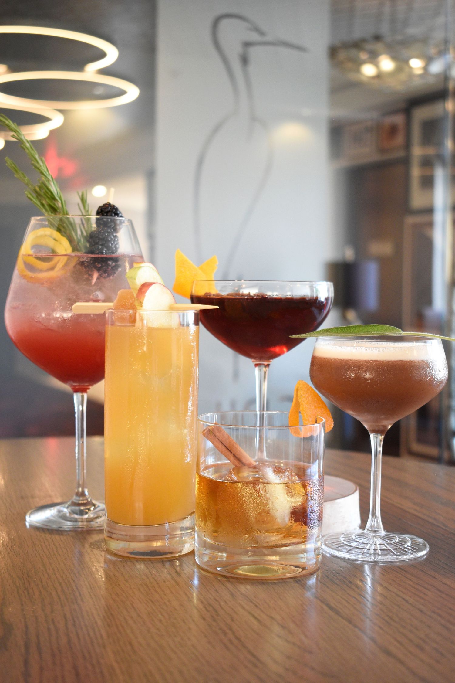 Five colorful craft cocktails garnished with fruits and herbs displayed on a wooden bar counter.
