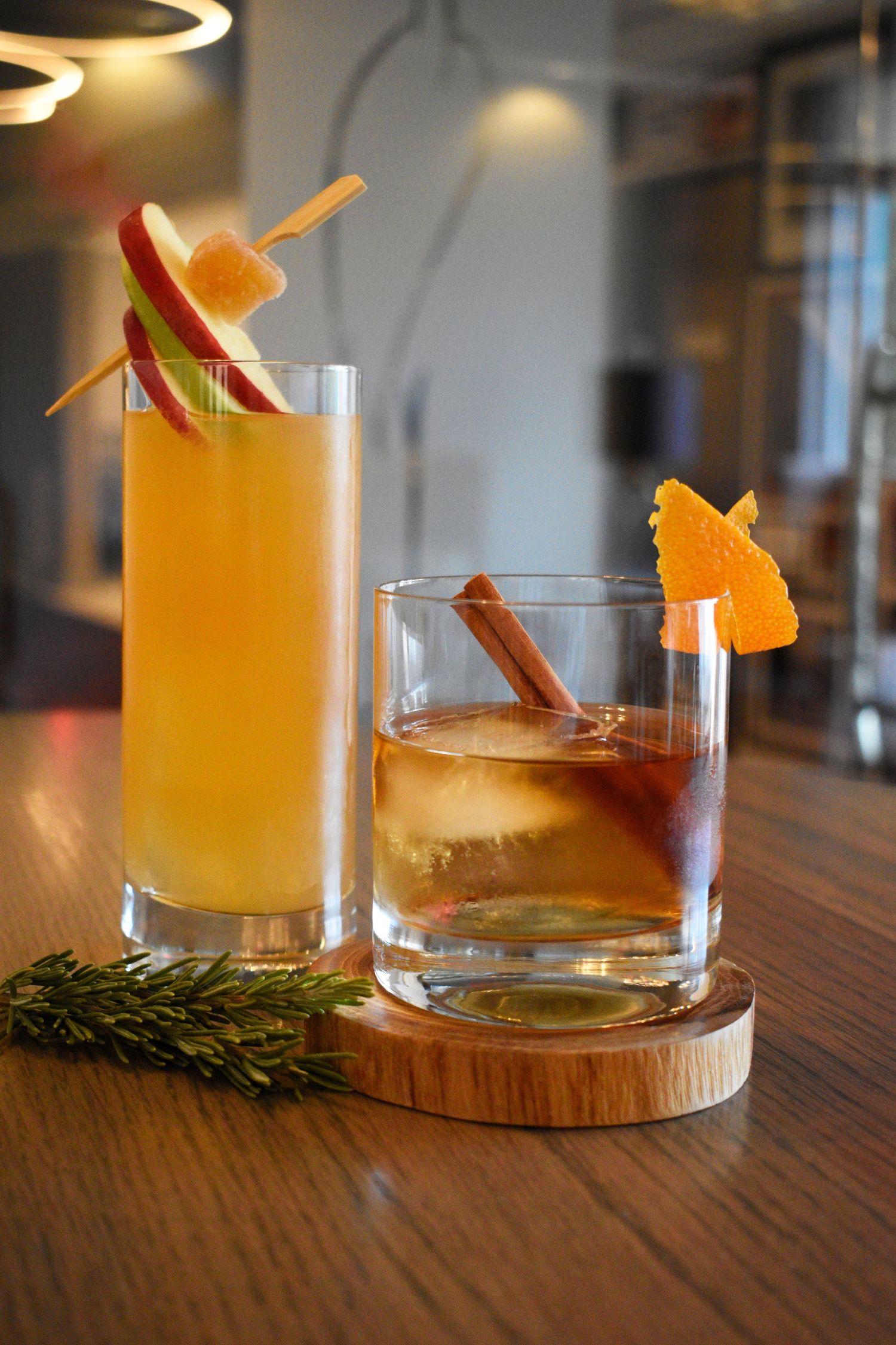 Two craft cocktails on a wooden bar, one tall orange drink with apple garnish and one rocks glass with cinnamon and orange peel.