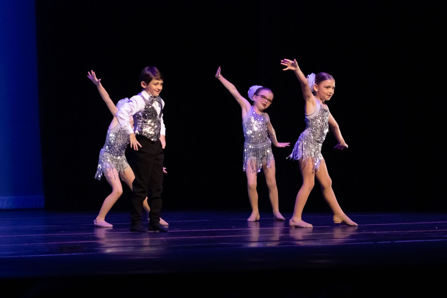 Four young dancers perform on stage in silver sequined costumes under blue stage lighting.