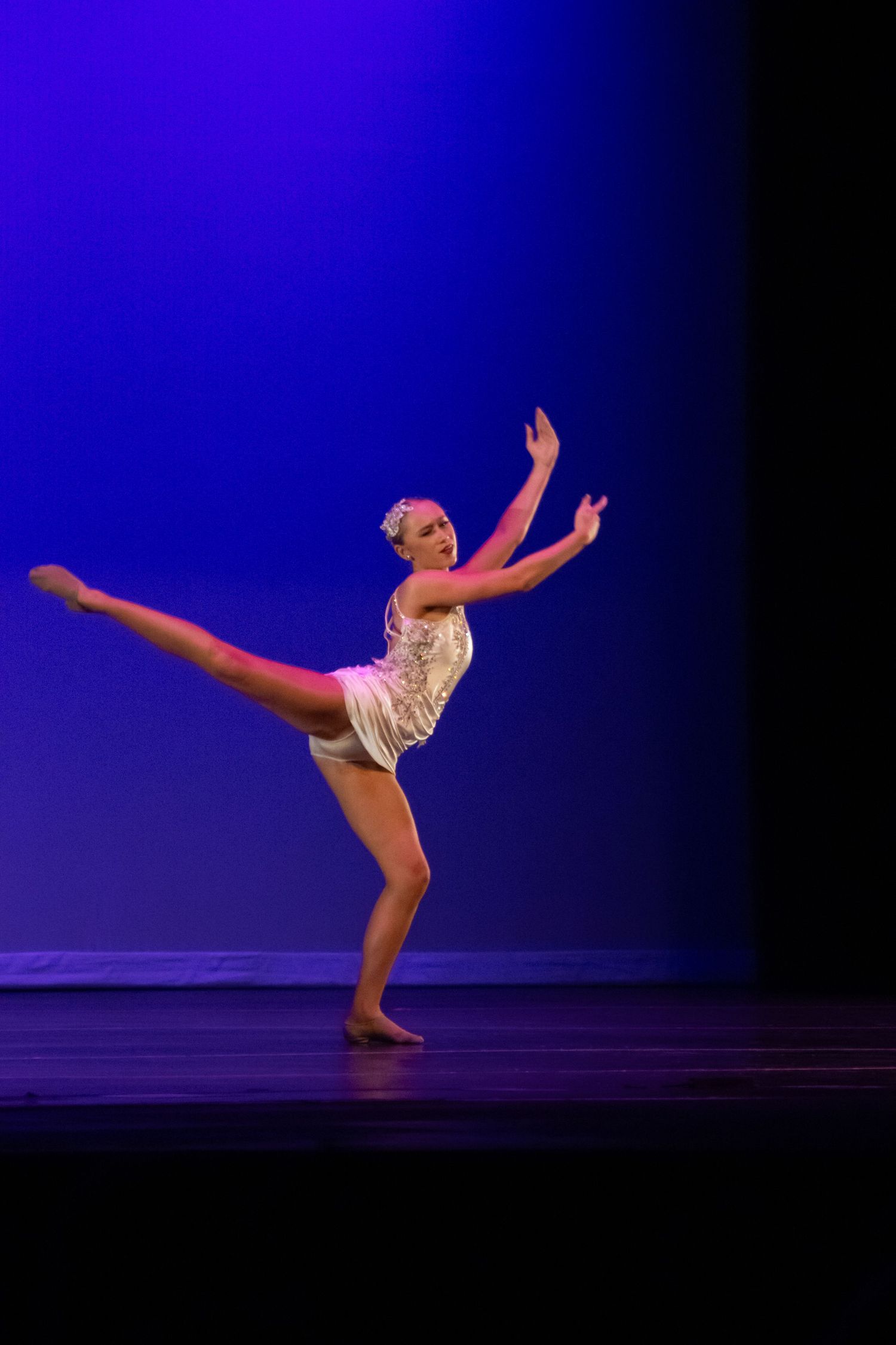 A female dancer performs an arabesque on stage in a white leotard under blue and pink stage lighting.