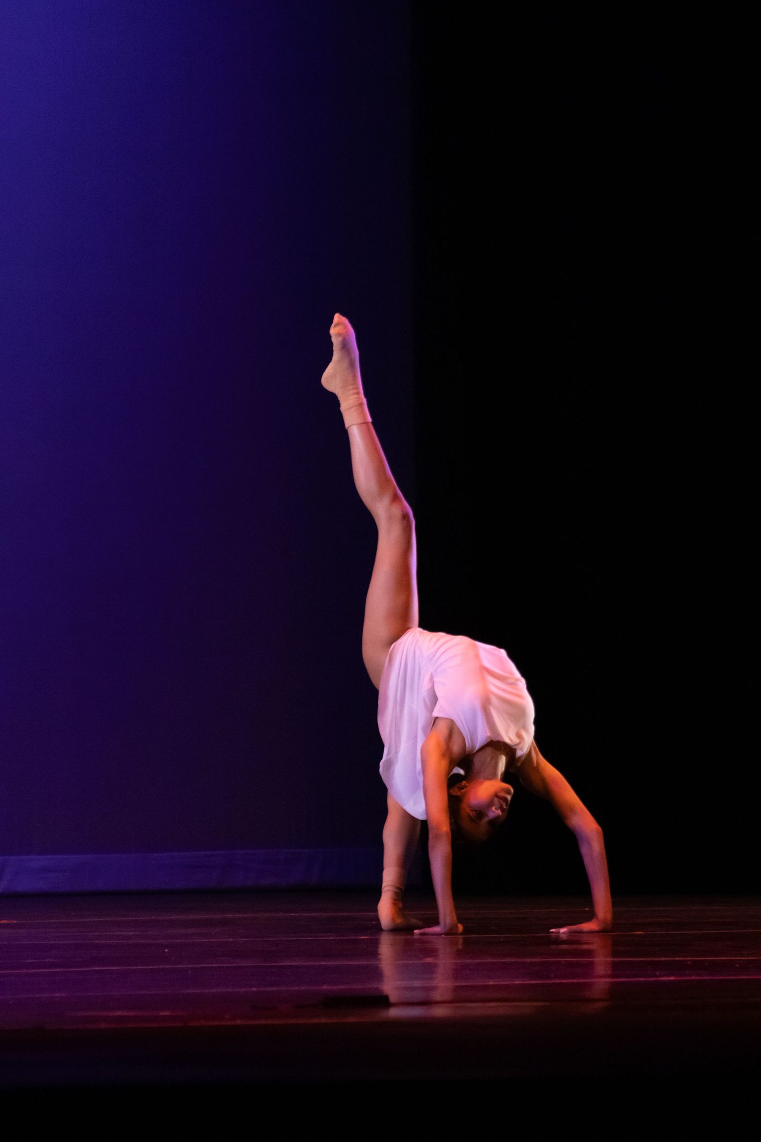 A young dancer performs a one-handed cartwheel with leg extended high on a darkened recital stage.
