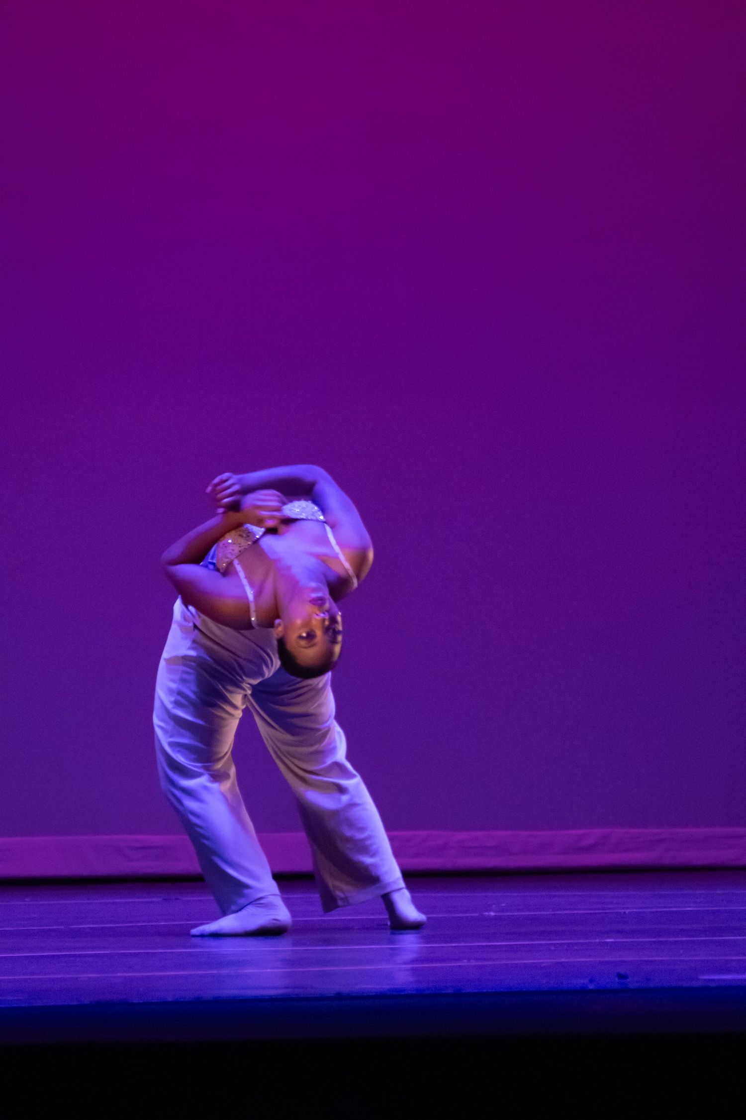 A contemporary dancer performs a deep backbend in white costume under purple stage lighting.