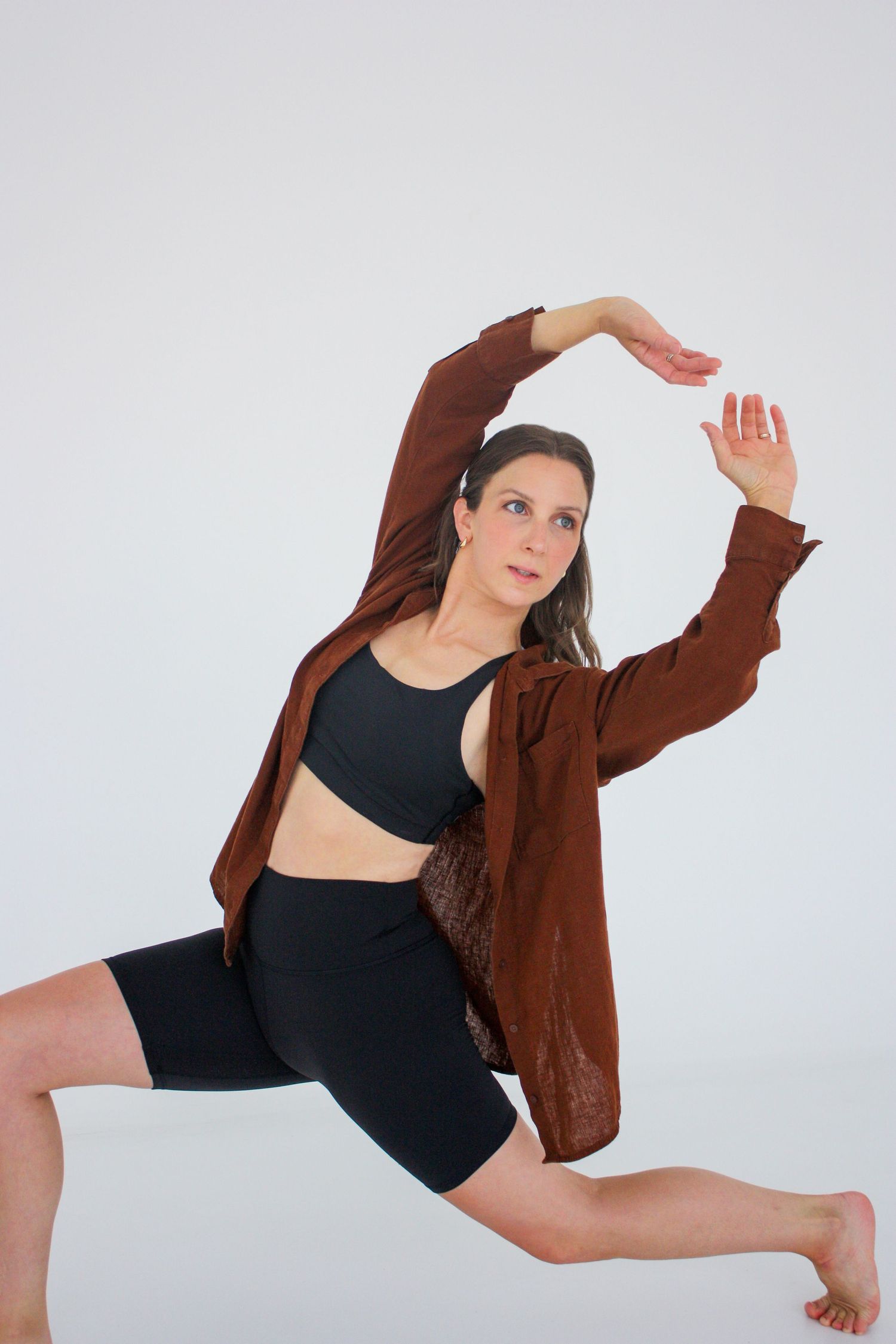 Female dancer in brown shirt and black shorts performs deep lunge with arched arms overhead. Branding photography Tampa, FL.