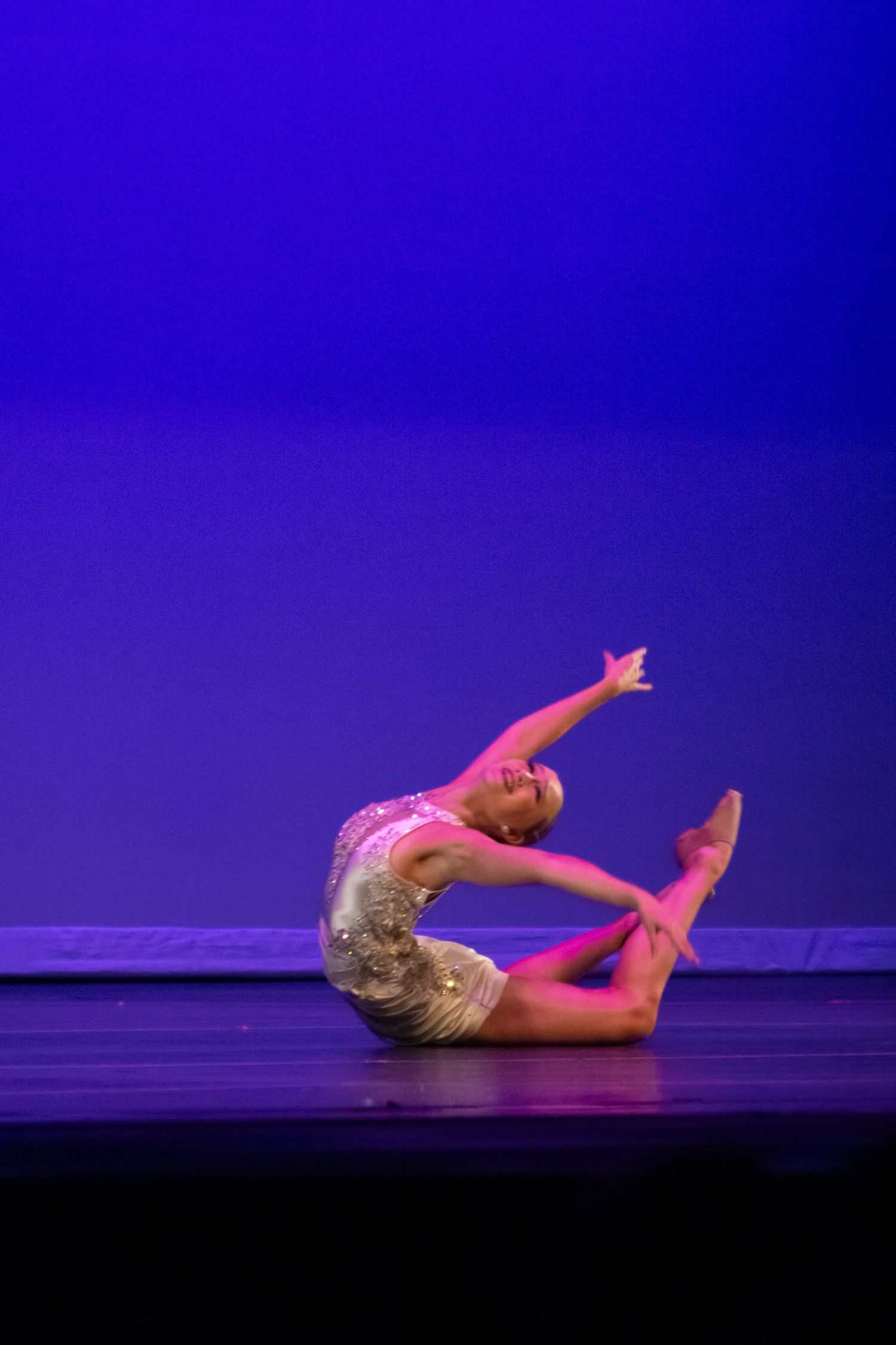 A flexible dancer in a pink leotard performs a stunning backbend on a blue-lit performance stage.