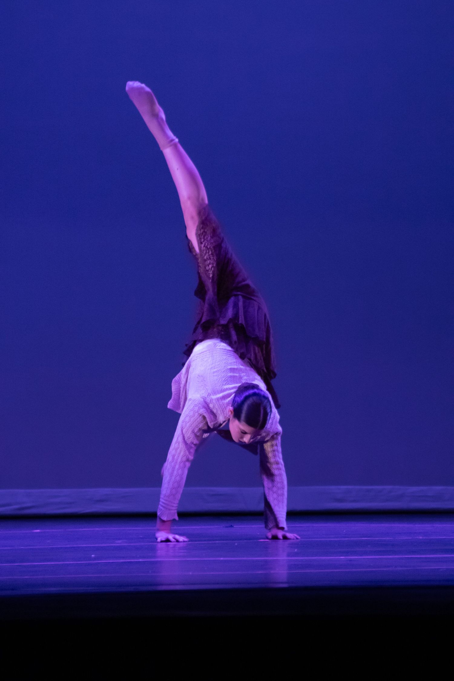 A dancer in white performs an inverted pose with one leg raised high on a blue-lit stage.