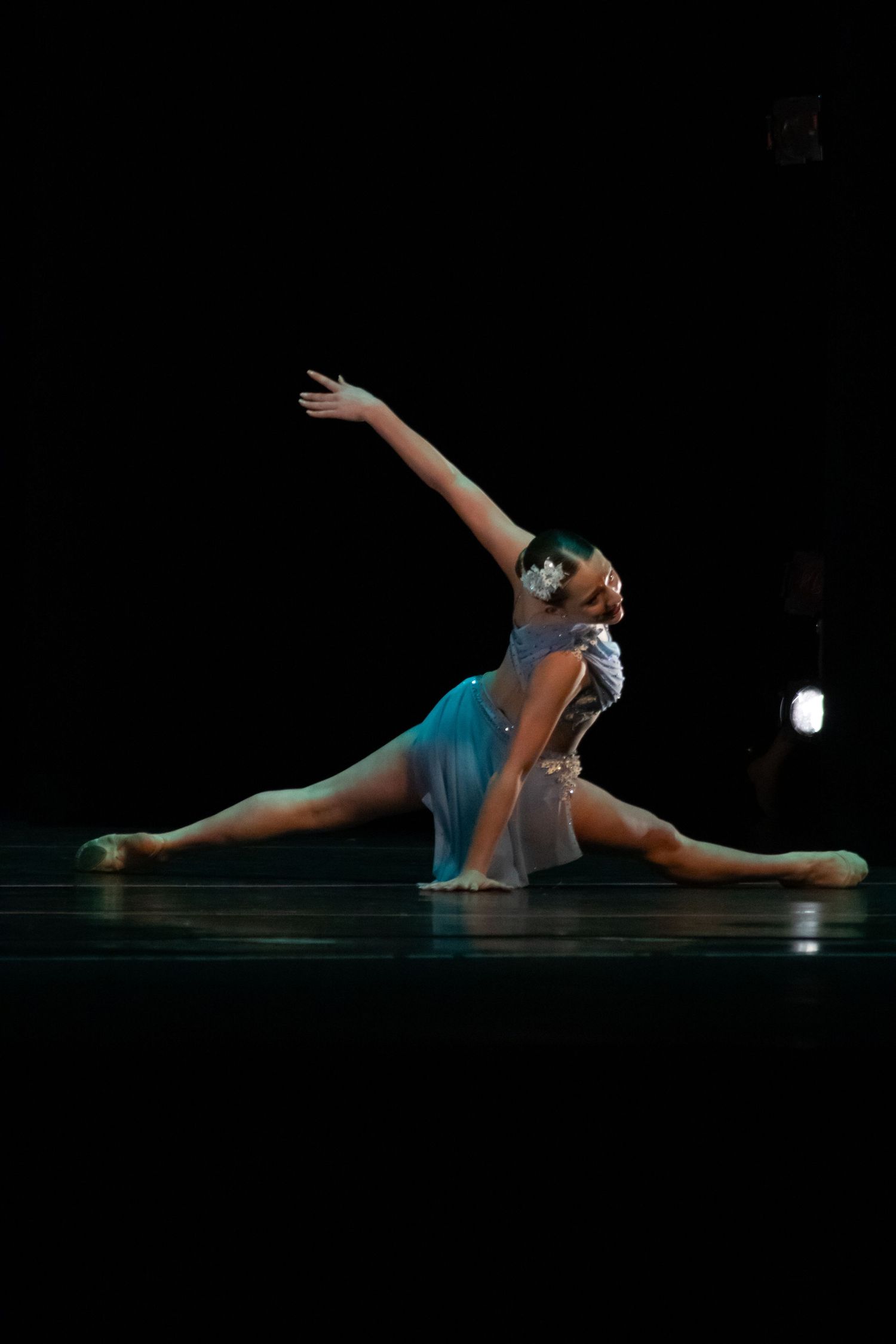 A dancer in a teal costume performs a side split with an elegant arm reach under dark stage lighting.