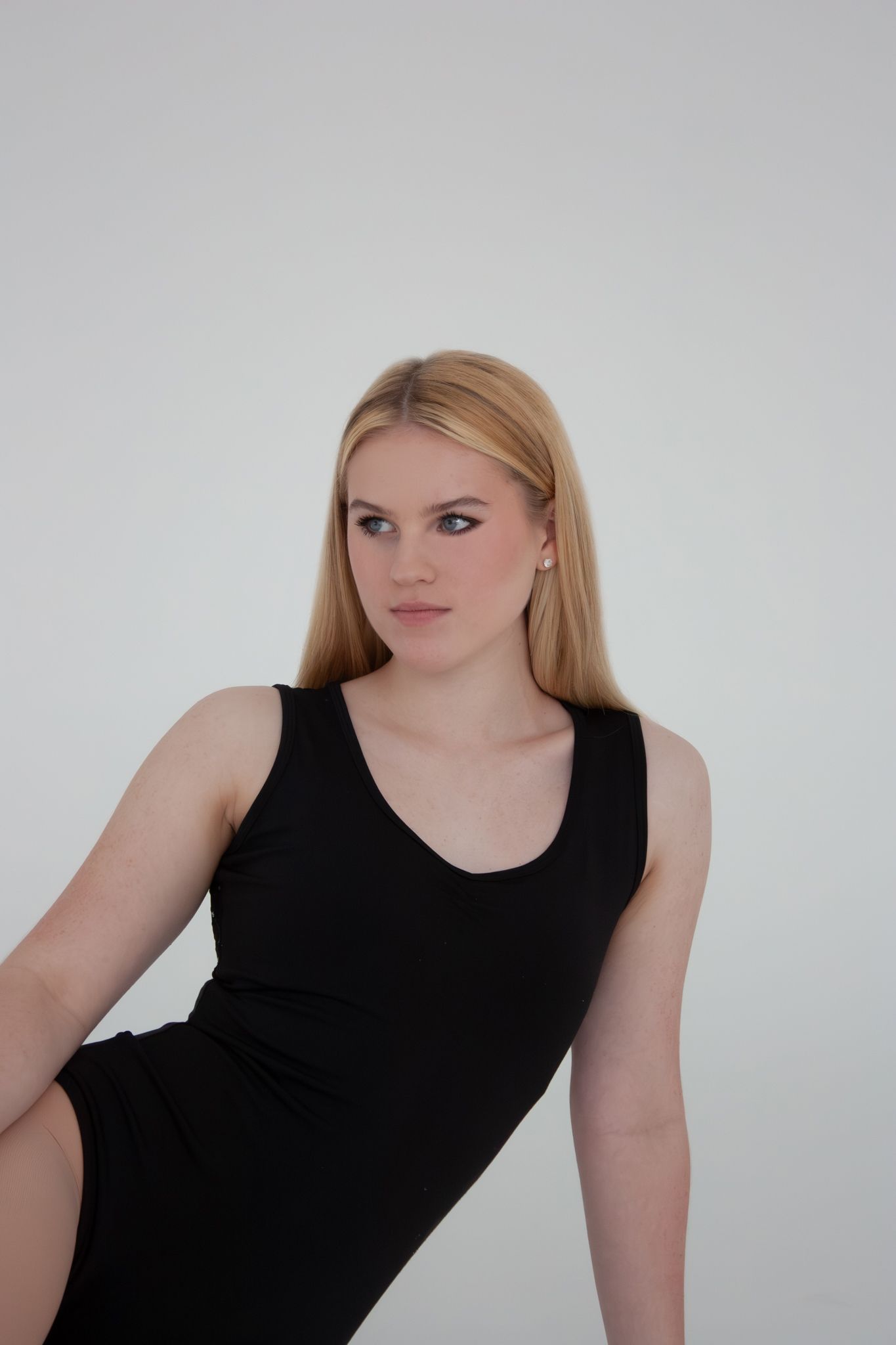 Blonde woman in black sleeveless dress sitting gracefully against white background.