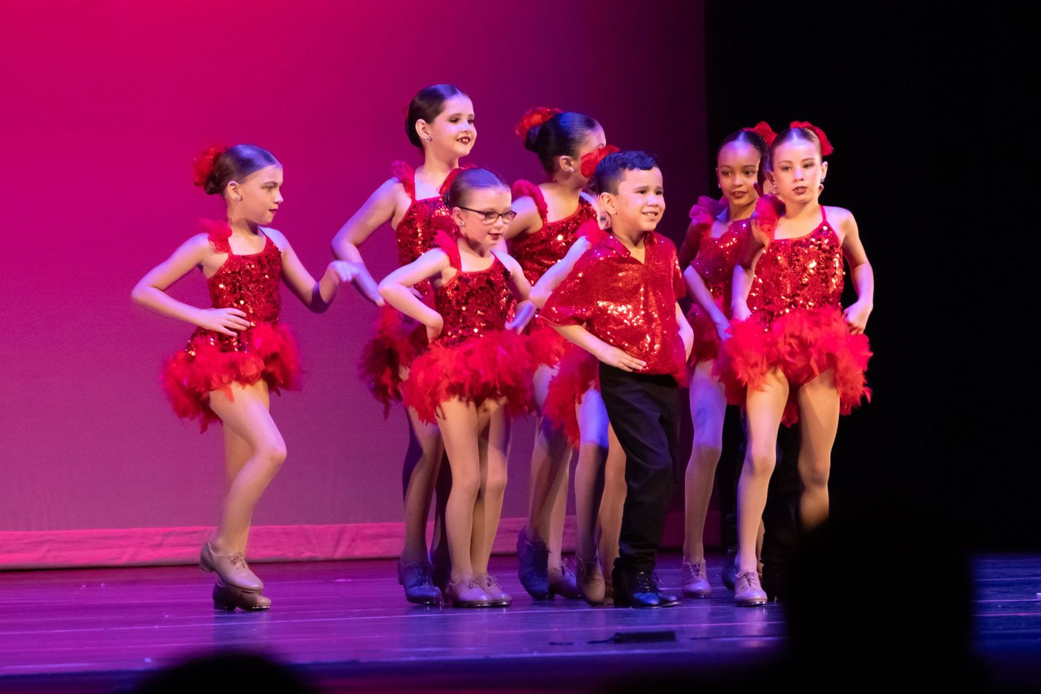 Children in red sequin costumes perform a dance recital on stage under bright lights.