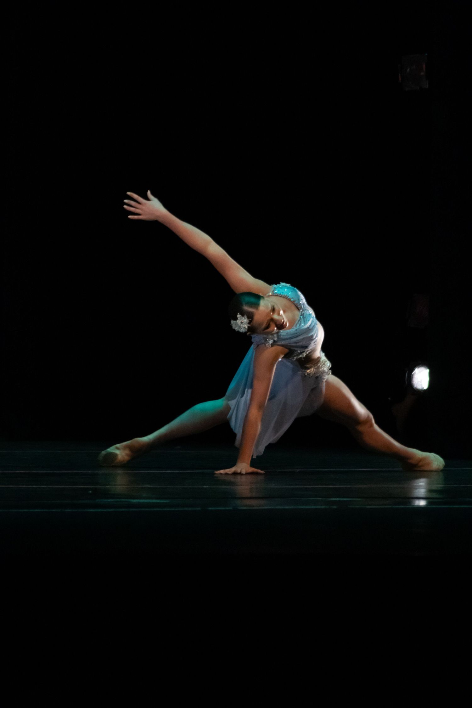 Dancer in teal costume performs elegant side lunge with arm extension on dark stage.