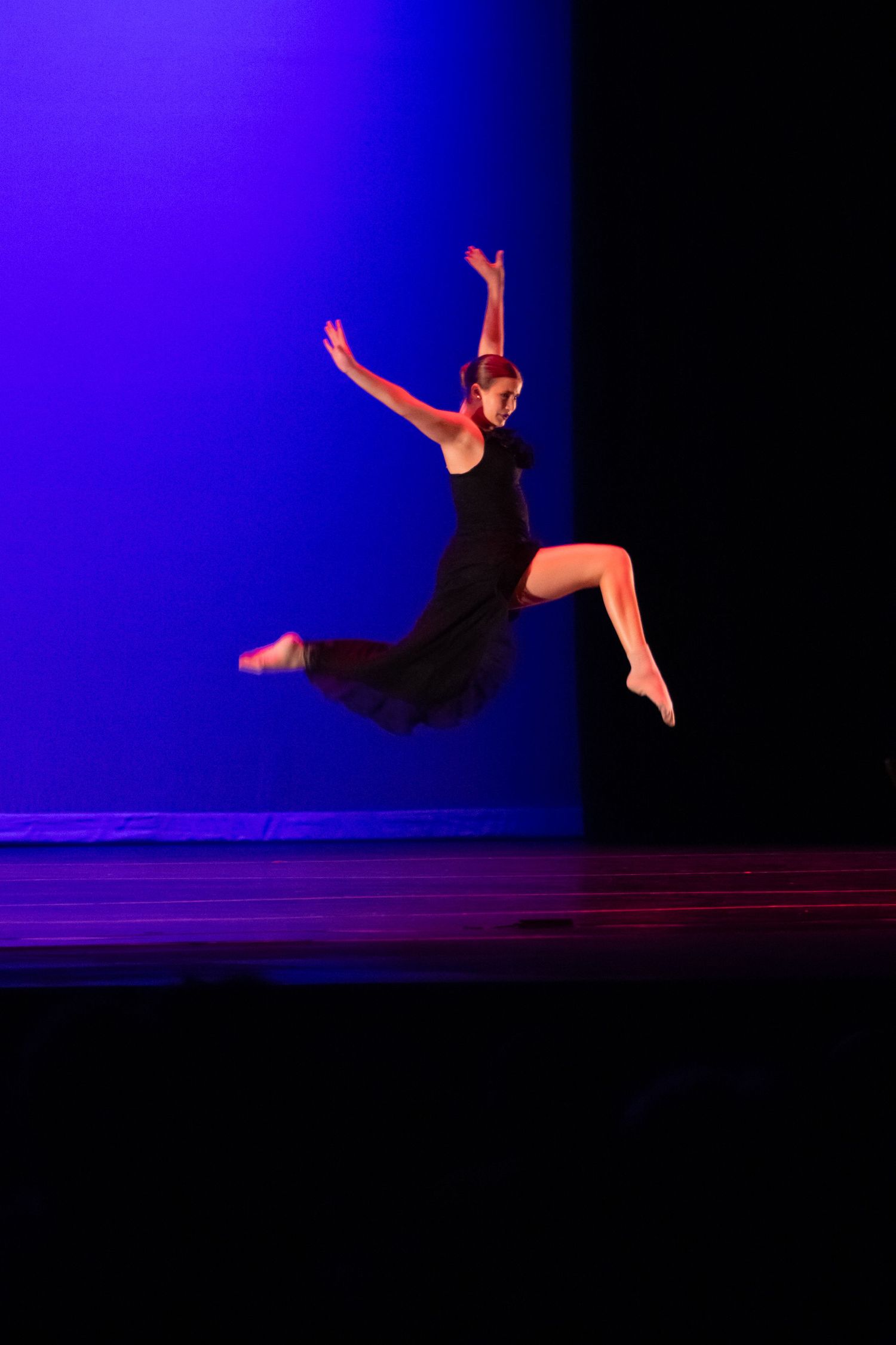 Female dancer in black dress leaps gracefully with arms raised on a blue-lit performance stage.