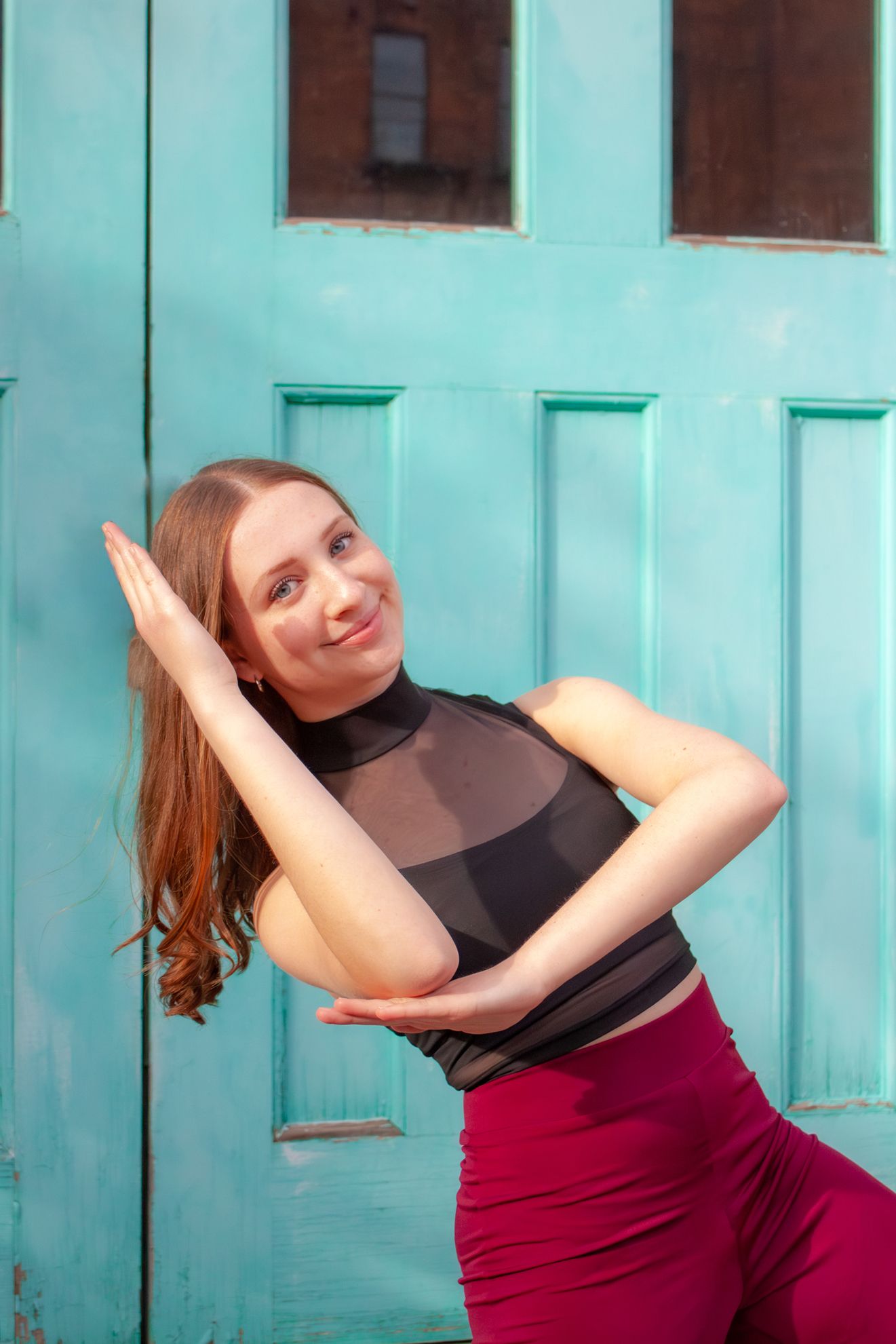 Female dancer in black top and red pants posing against a turquoise door smiling.