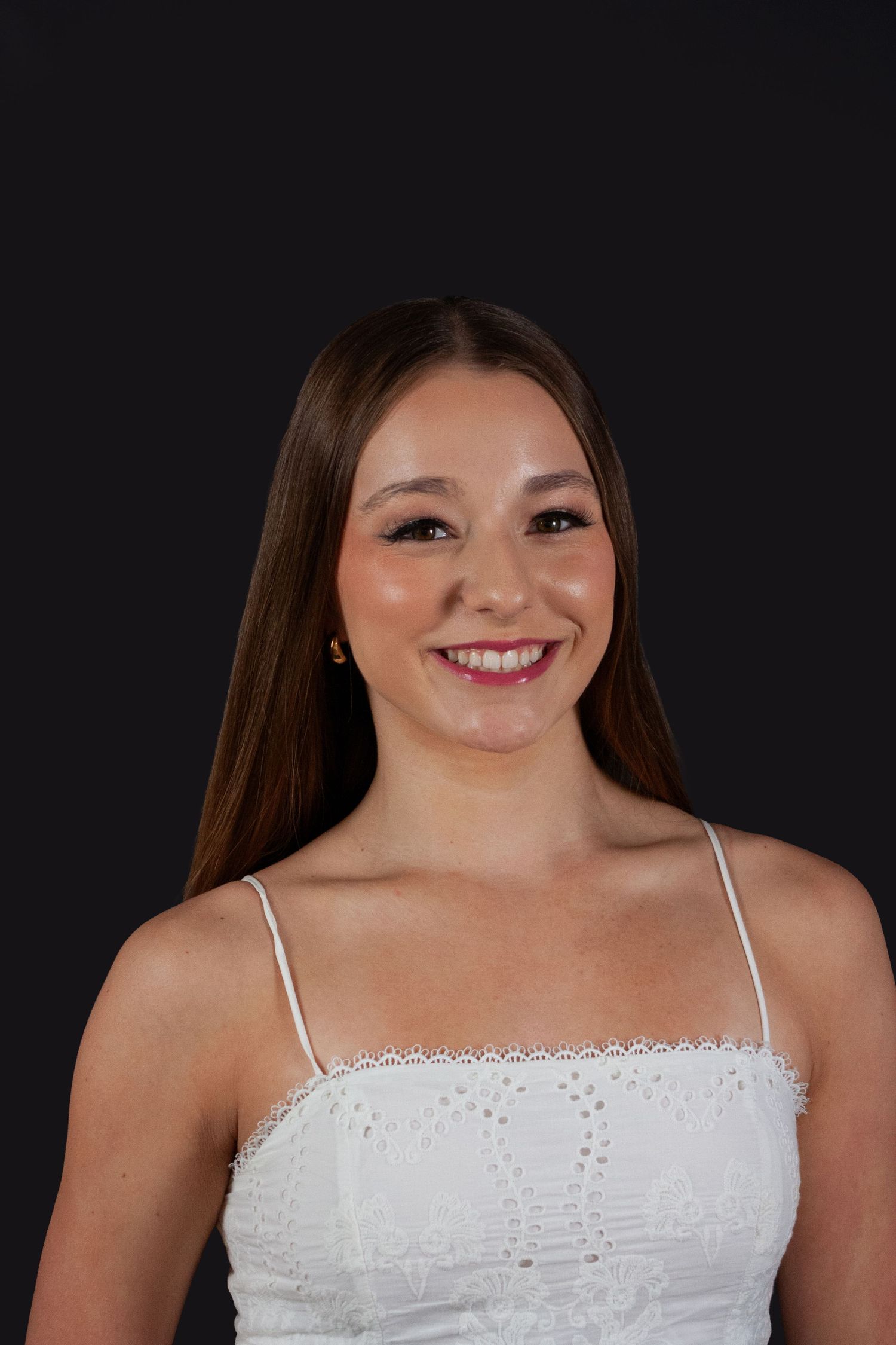 Smiling young woman with long brown hair wearing white lace spaghetti strap top against black background.