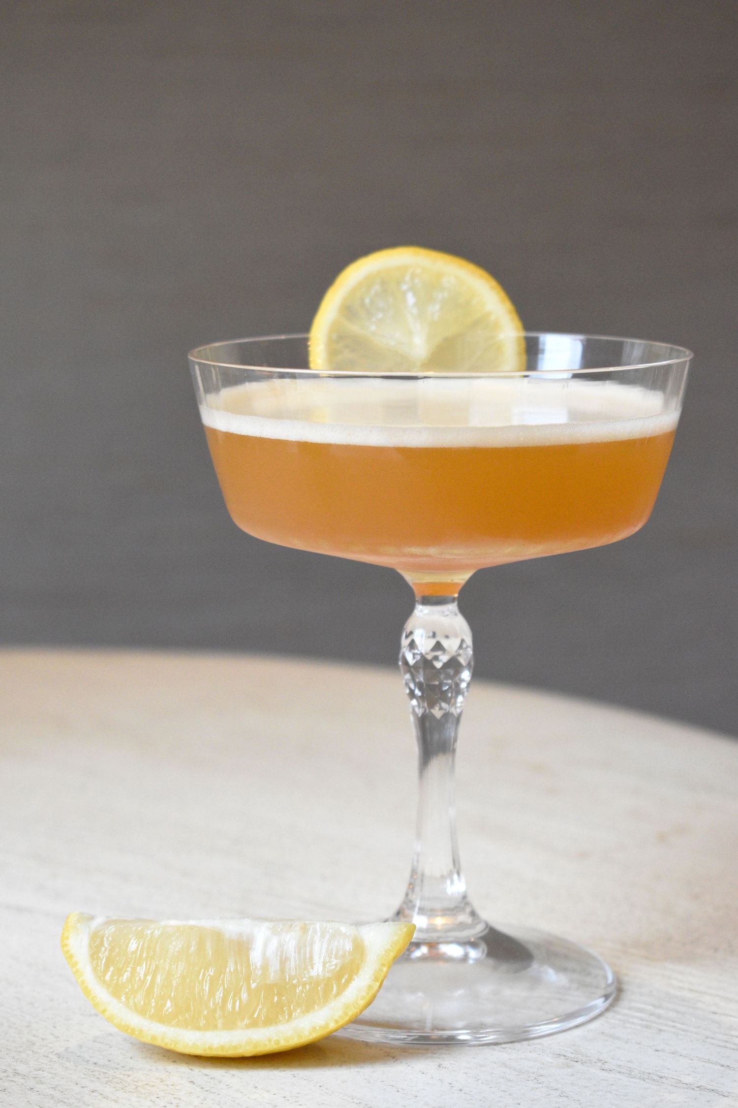 Elegant amber cocktail in a crystal coupe glass garnished with a lemon wheel and slice on a light wooden surface.