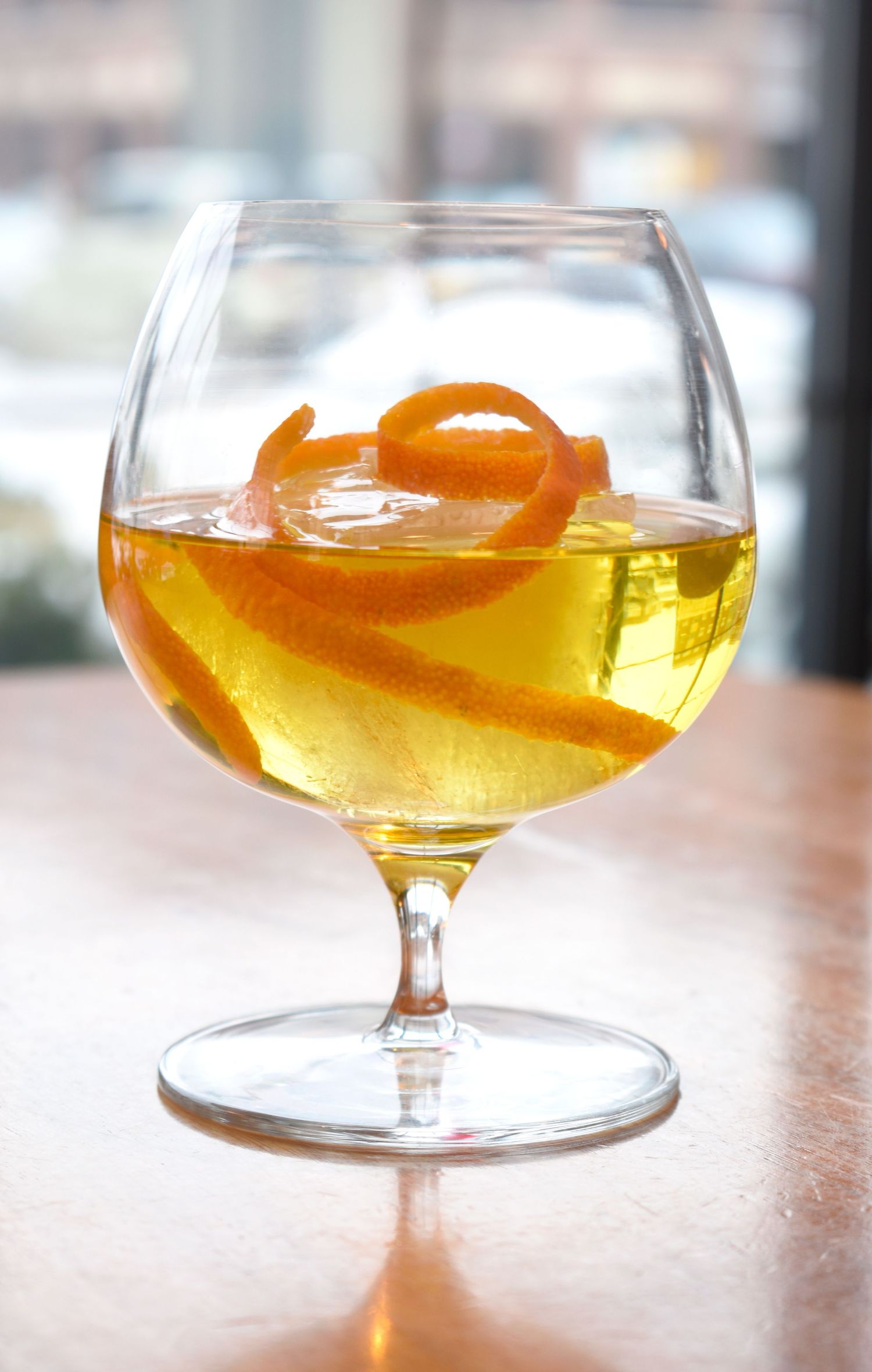 A golden cocktail with a curled orange peel garnish served in a large brandy snifter glass.