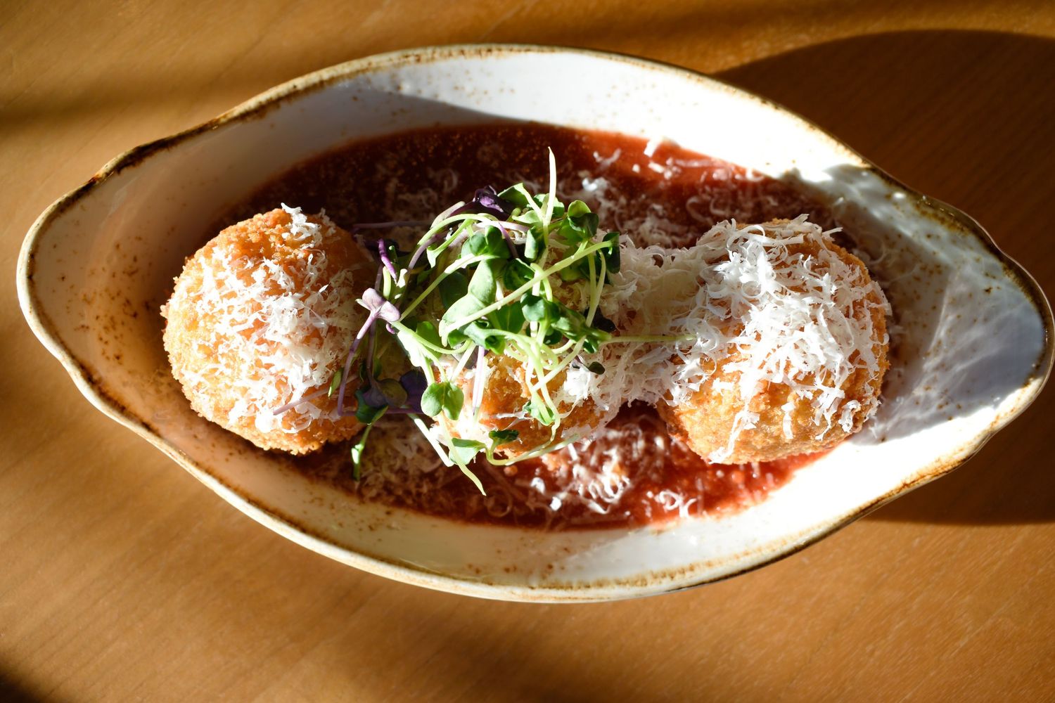 Two golden fried arancini rice balls in marinara sauce topped with shaved parmesan and microgreens.