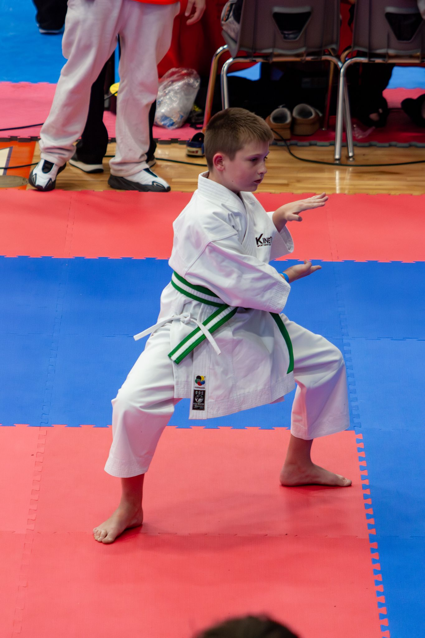 A young boy in a white karate gi with green belt performs a kata stance on a competition mat.