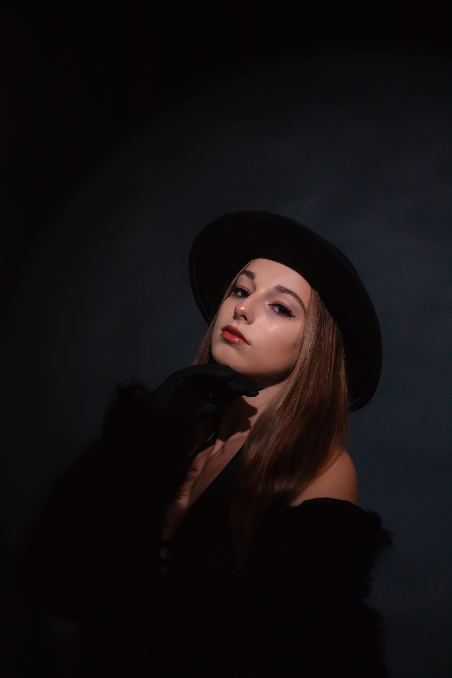 Young woman in black wide-brim hat and fur jacket poses in dark atmospheric studio portrait.
