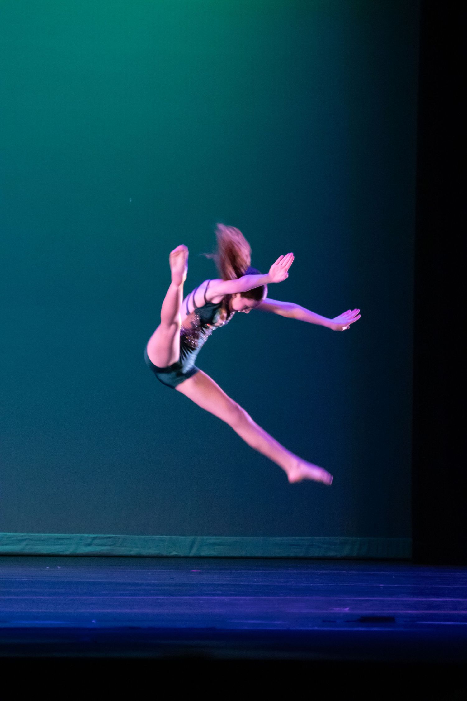 A dancer leaps mid-air in a dynamic jump pose on a teal-lit performance stage.