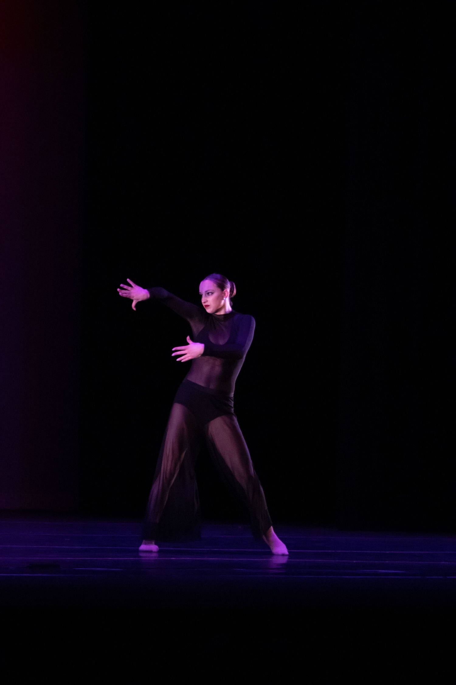 A dancer in an all-black sheer costume performs a dramatic contemporary solo on a darkened stage.