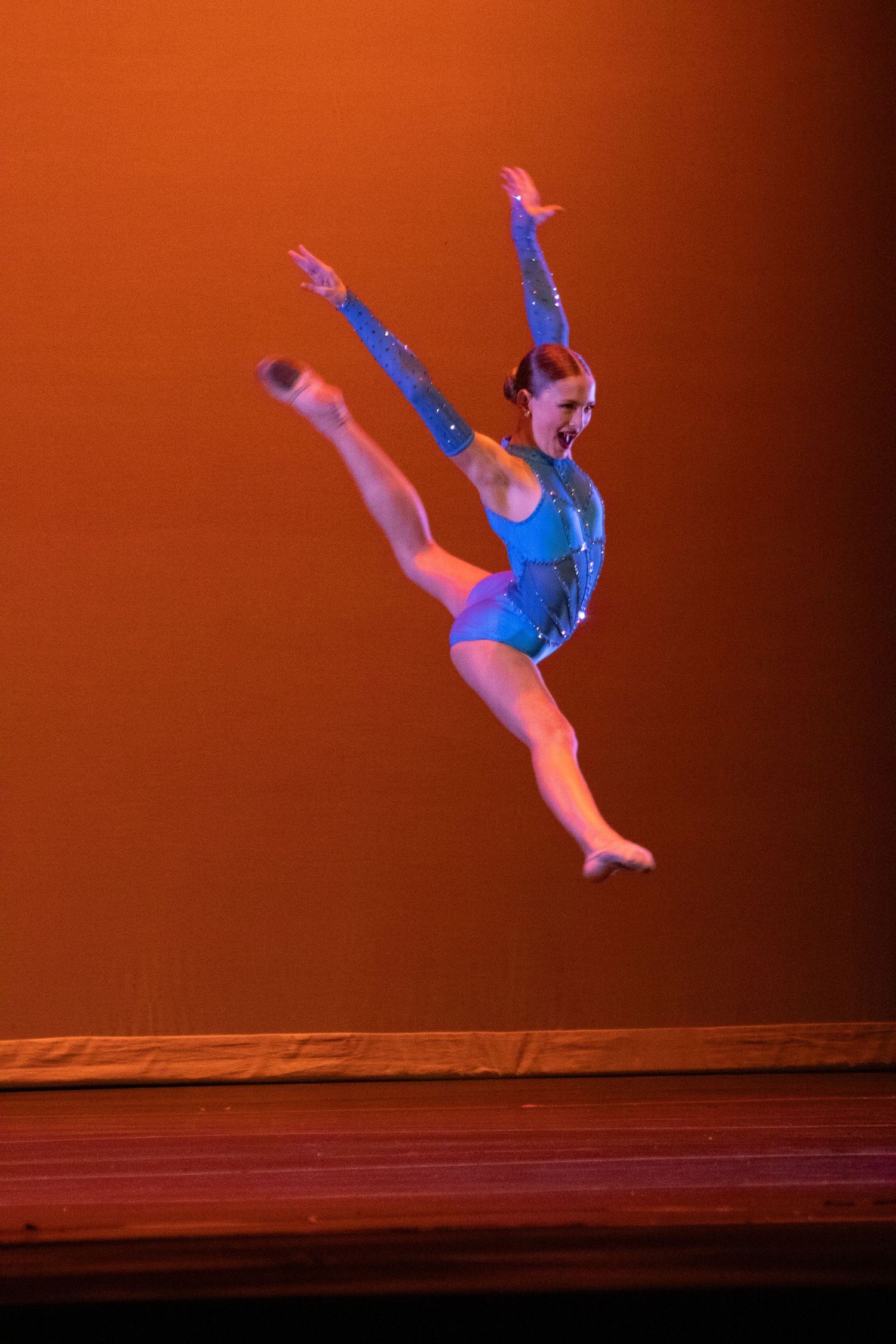 A gymnast in a blue leotard leaps high on a warmly lit stage.