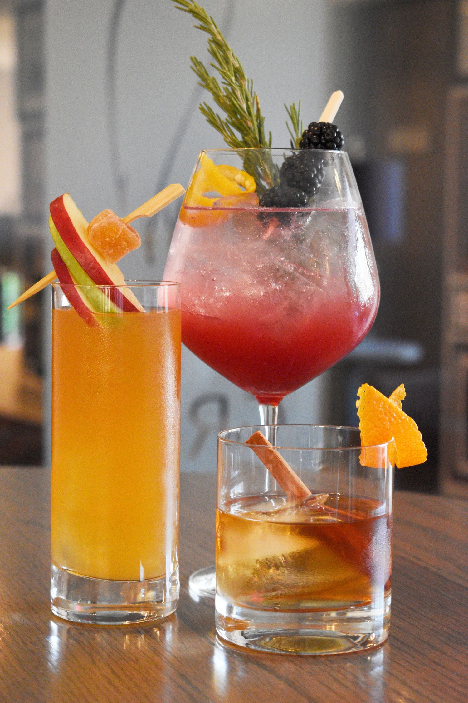 Three colorful cocktails garnished with fresh fruit, rosemary, and blackberries displayed on a wooden bar.