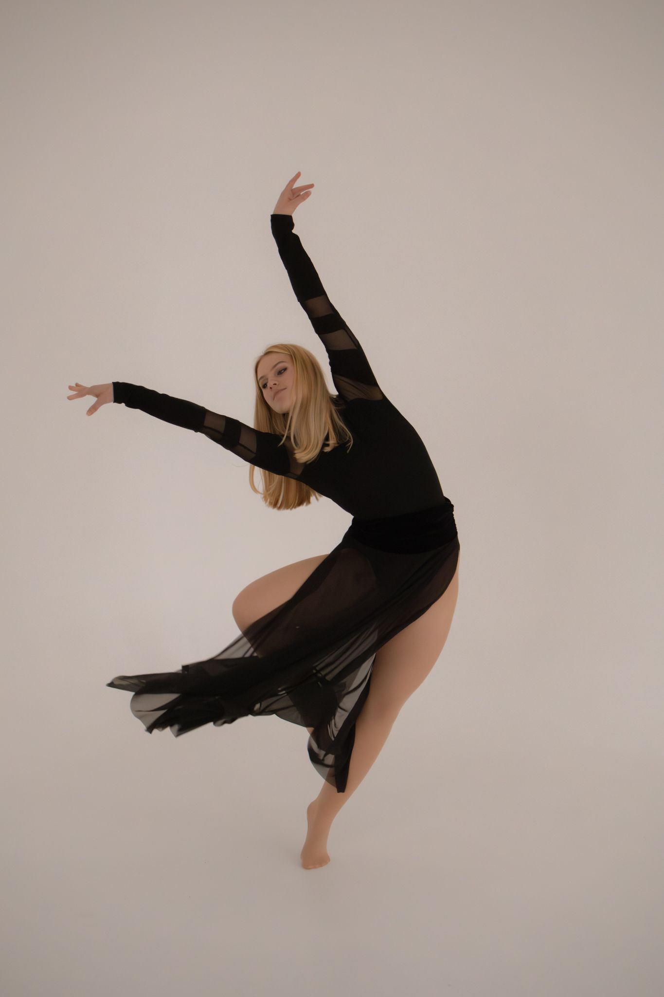 Blonde dancer in sheer black dress mid-leap with arms raised against white background.