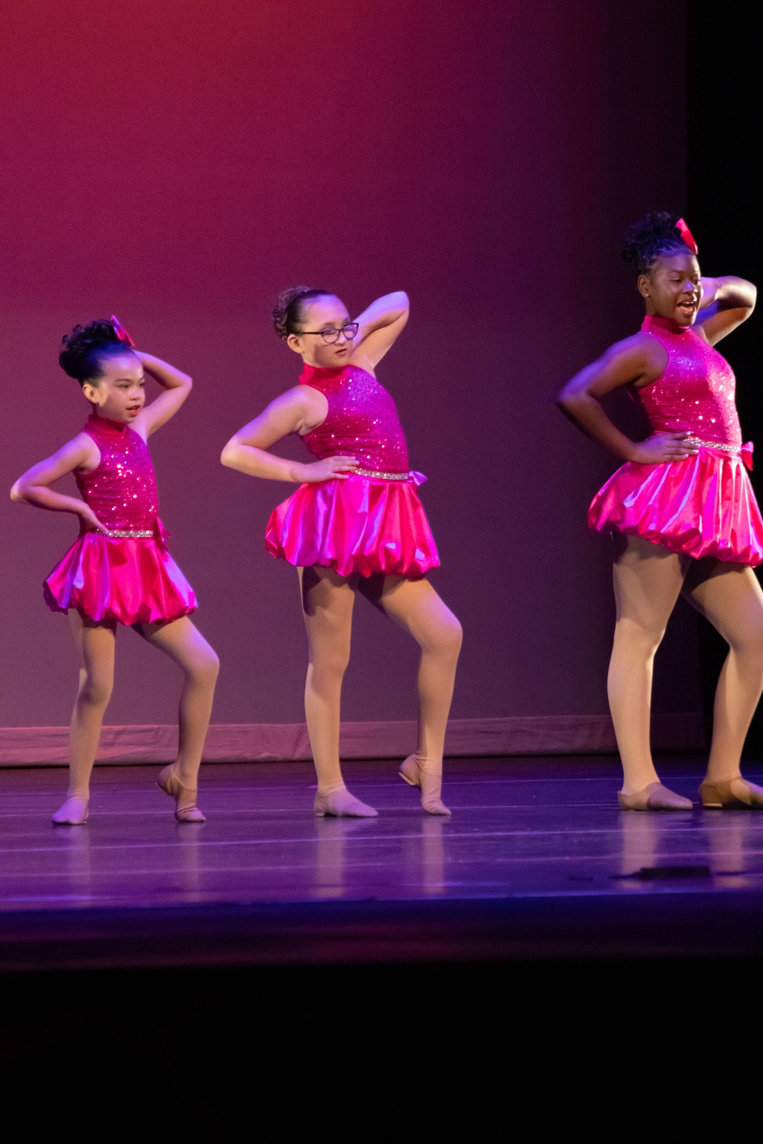 Three girls in pink sequin costumes perform an energetic dance routine on a lit stage.